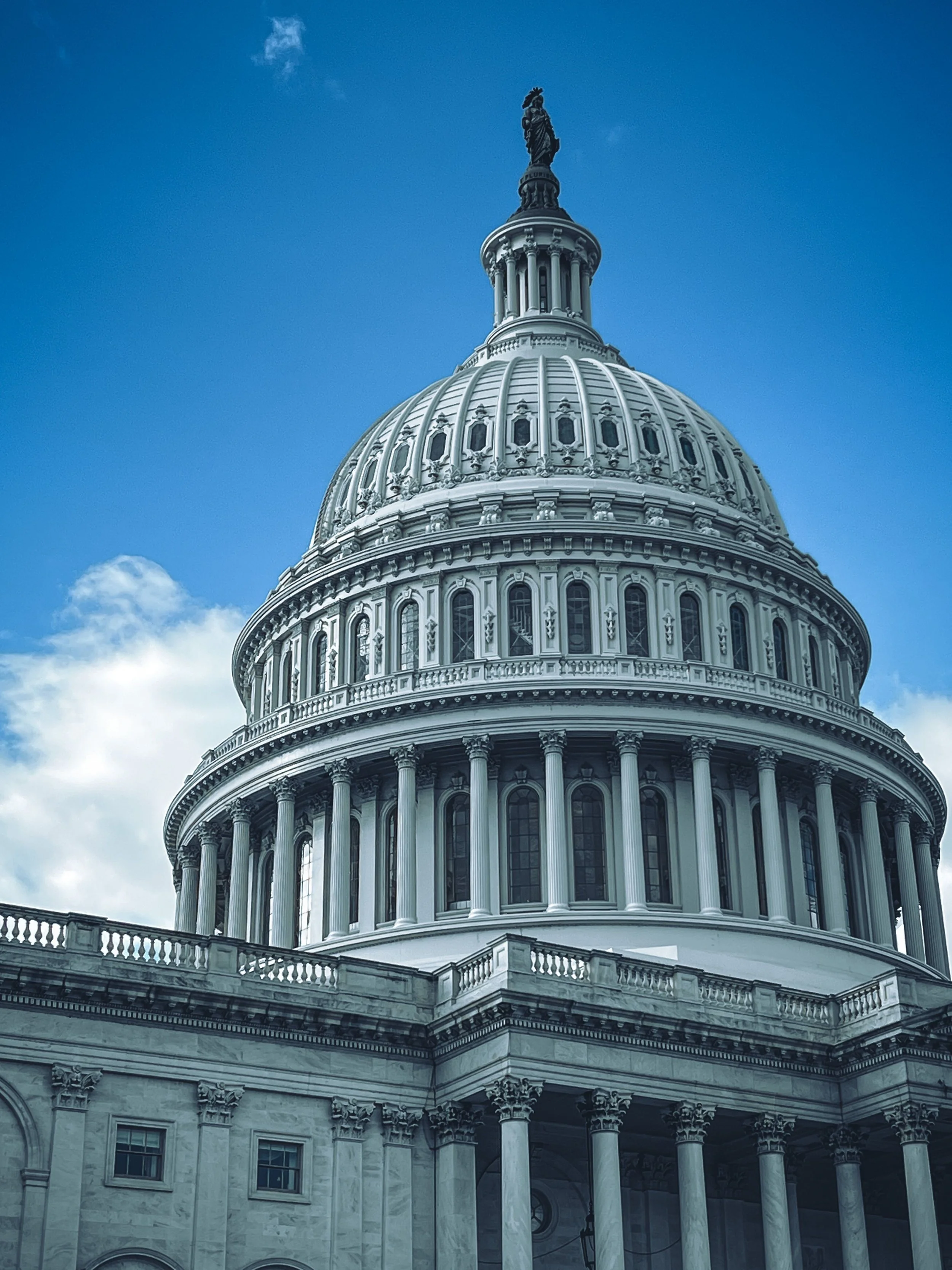 The United States Capitol building with its iconic dome, detailed architecture, and a statue on top, under a partly cloudy blue sky.