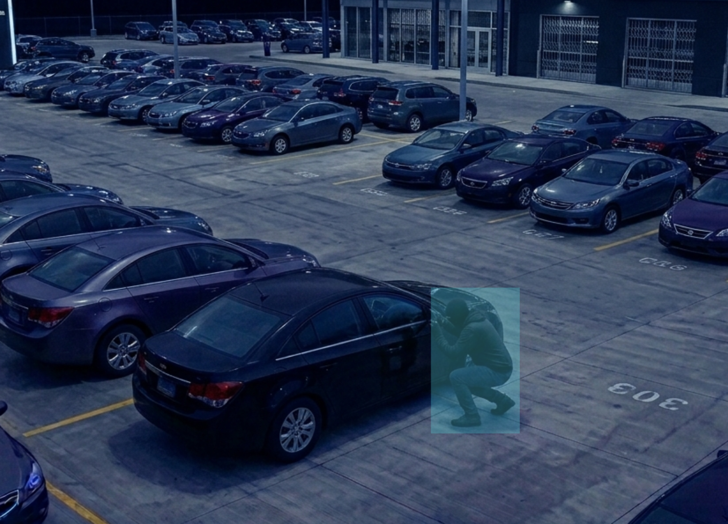 A person crouching next to a car in a dark parking lot at night attempting to break in, highlighted in a blue rectangle indicating surveillance detection.