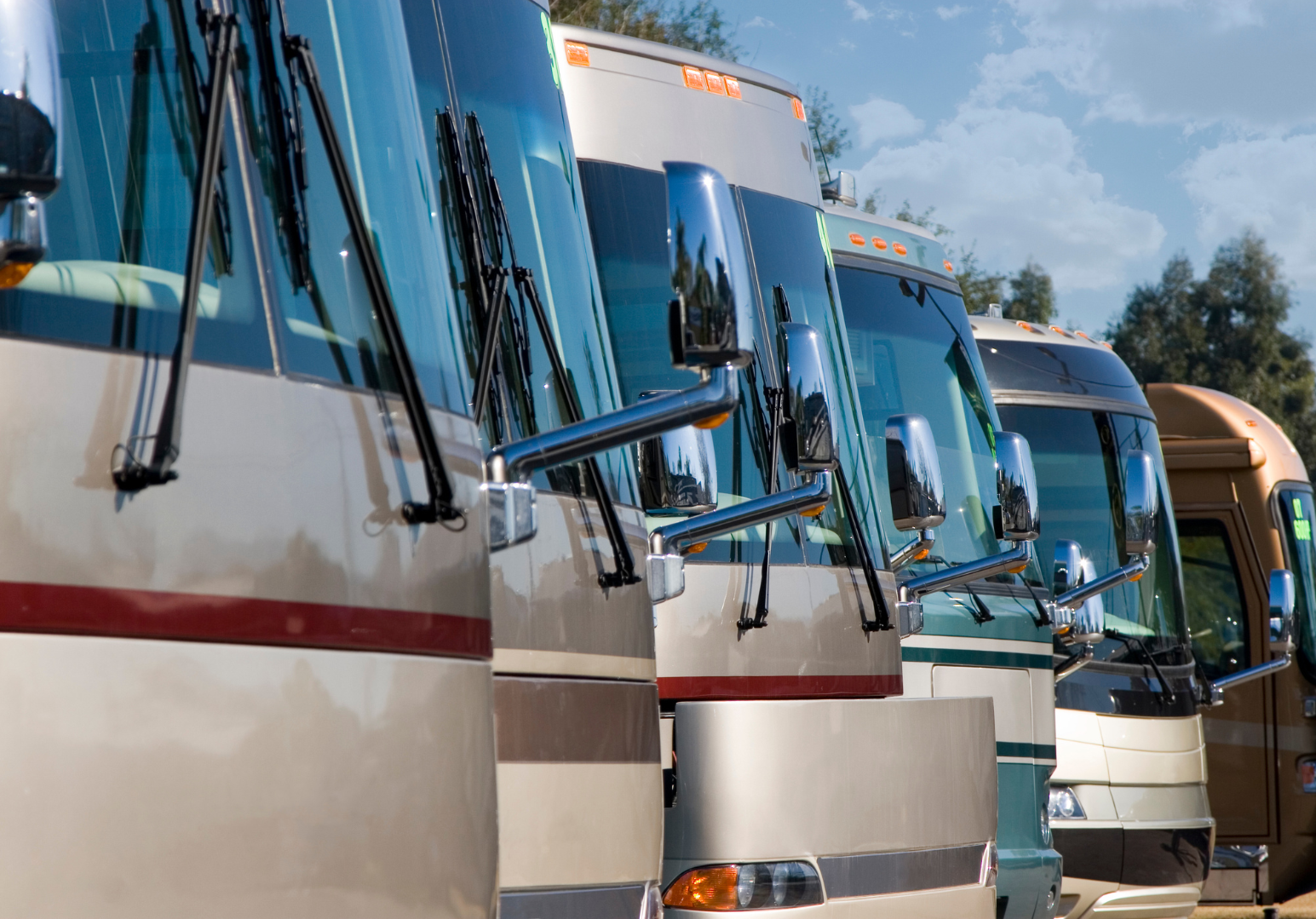 Multiple recreational vehicles (RVs) parked side by side outdoors against a blue sky with some clouds, trees in the background.