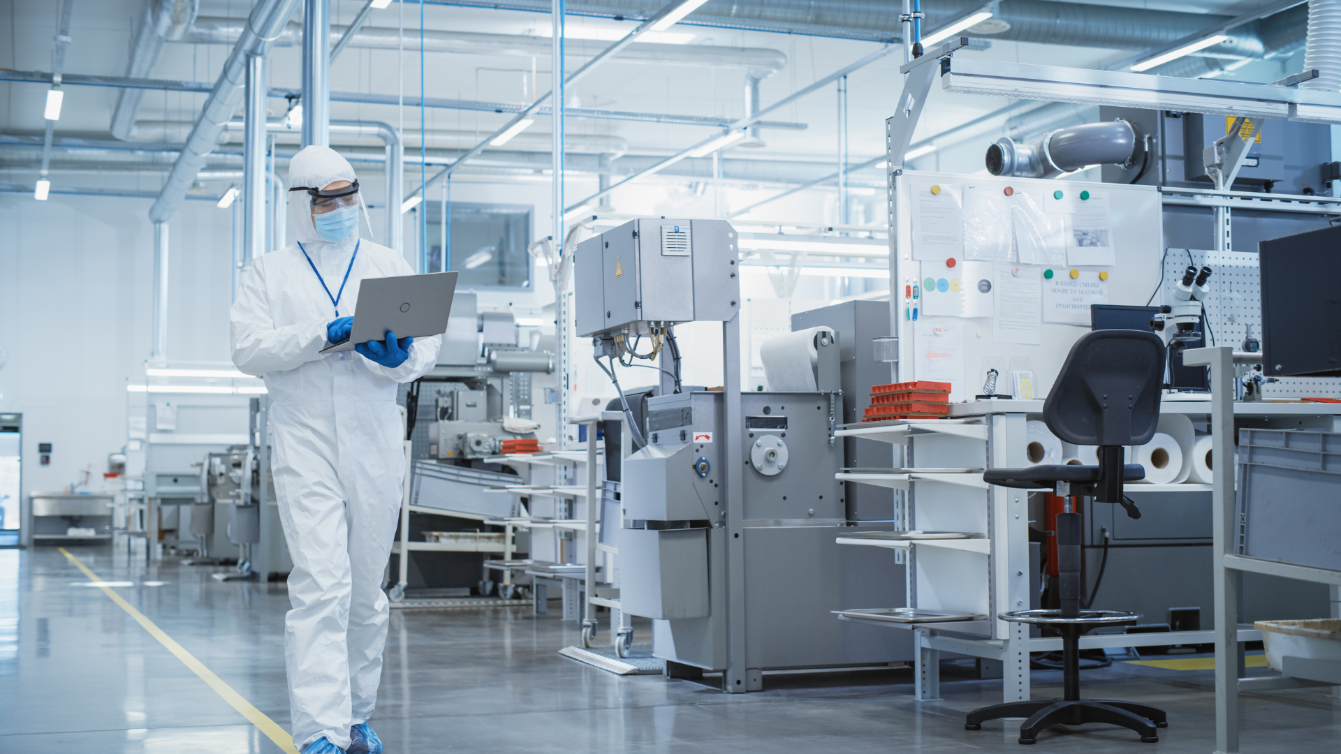 A scientist in protective gear, including a white suit, face mask, gloves, and glasses, working in a modern laboratory with various equipment, machinery, and workstations.