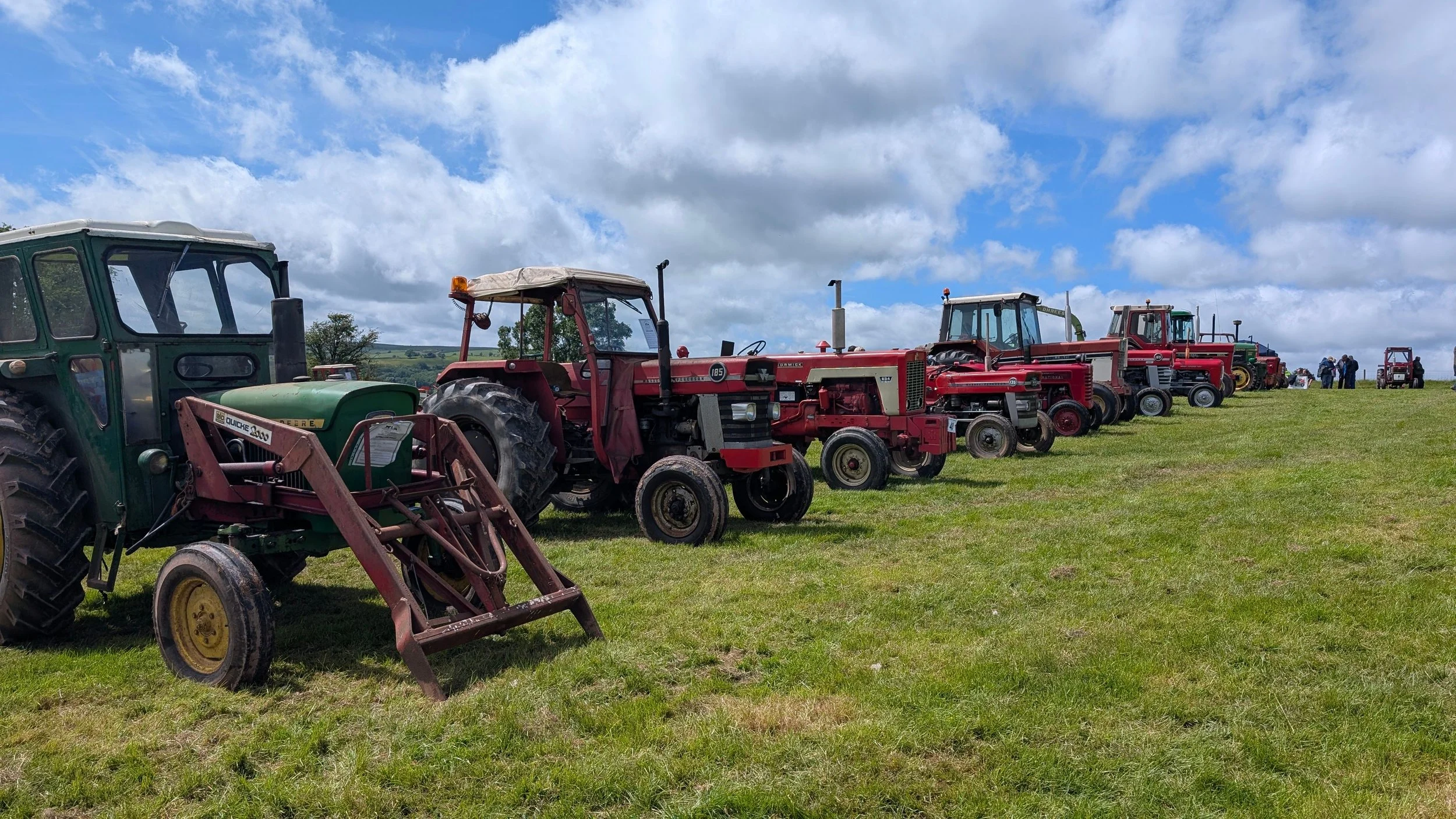 Row of vintage tractors lined up on a grassy field under a partly cloudy sky during daytime.