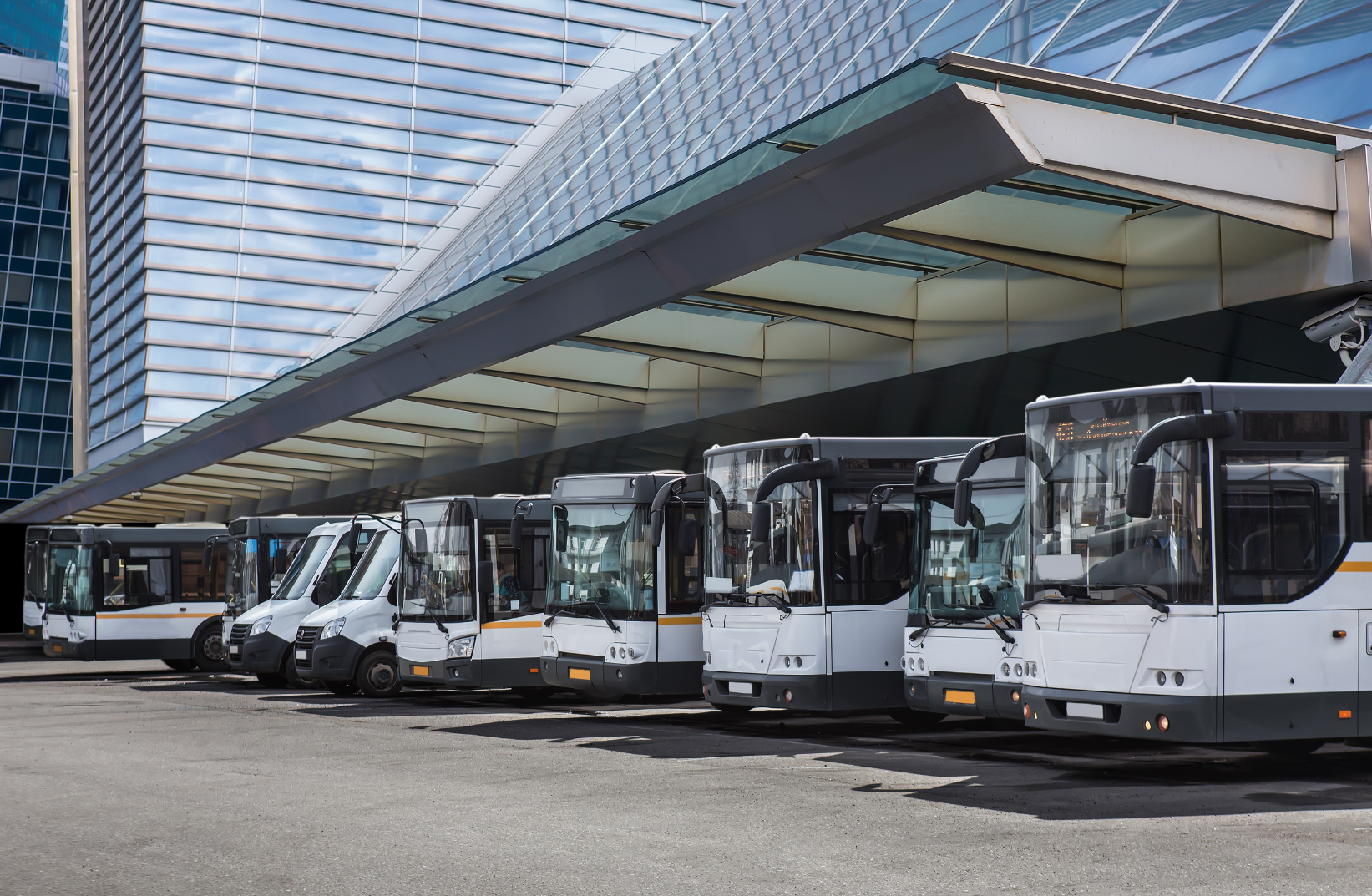Multiple white and black city buses parked in front of a modern glass building with a canopy.