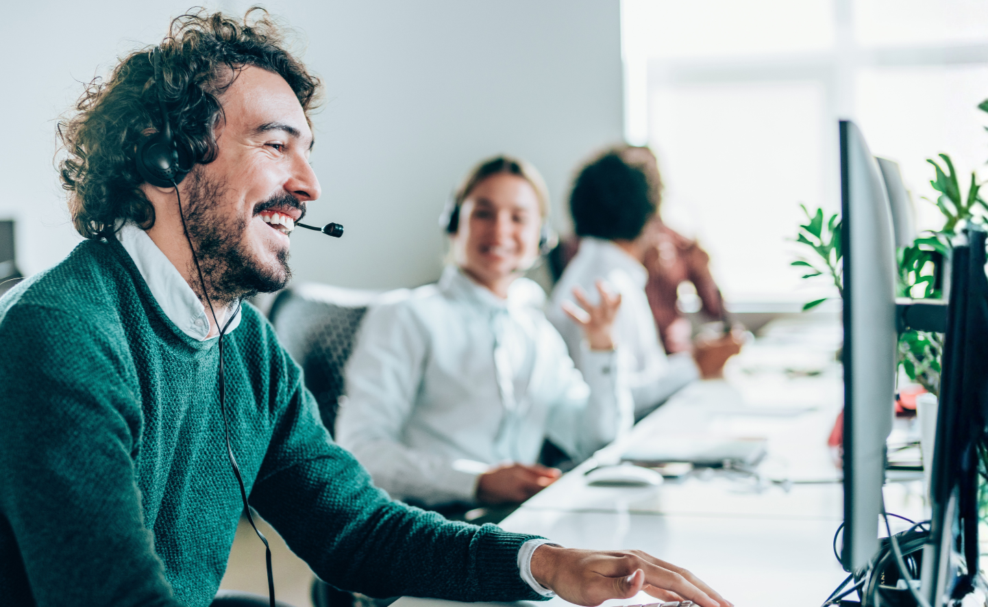 A man with curly hair and a beard wearing a headset, smiling while working at a computer in an office, with two colleagues in the background also using headsets and working at their desks.