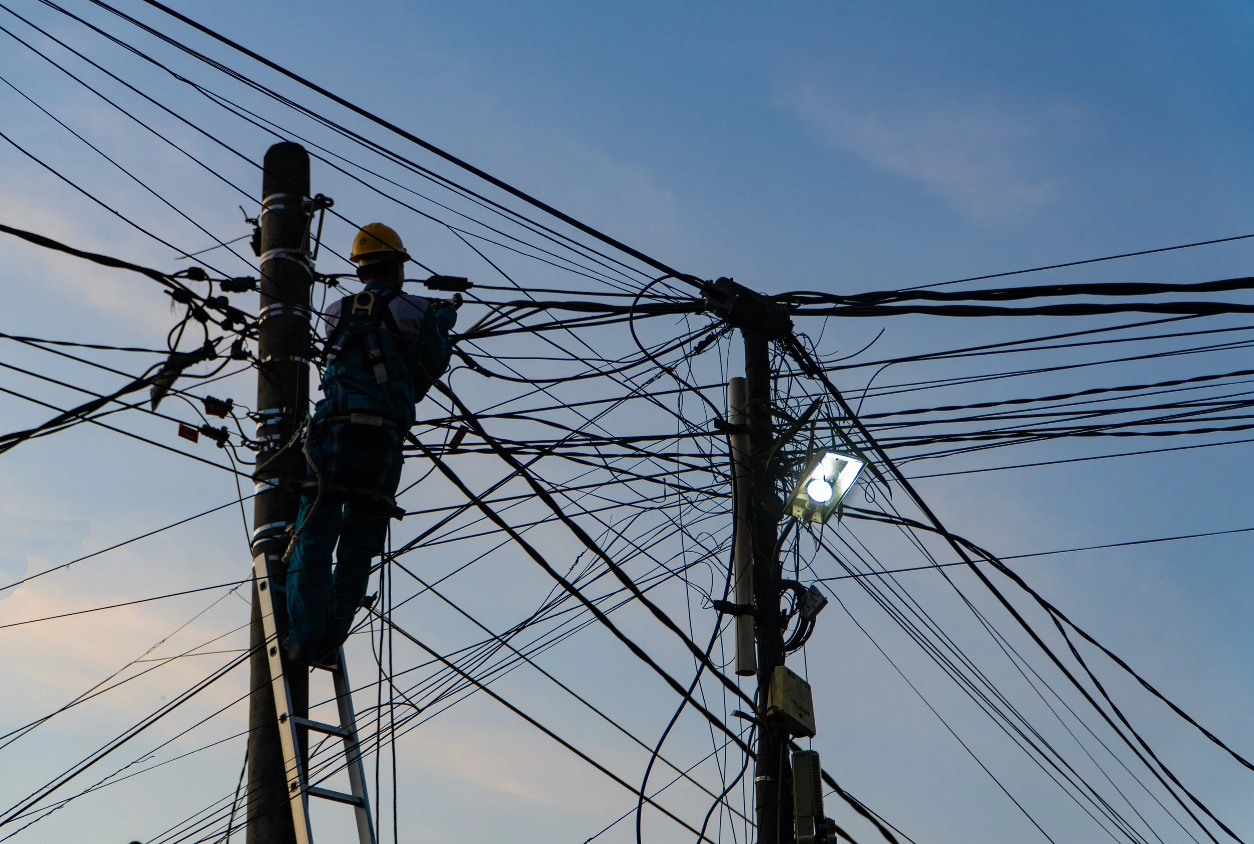 A worker wearing a yellow safety helmet and harness is working on an electric utility pole with multiple power lines against a clear blue sky.