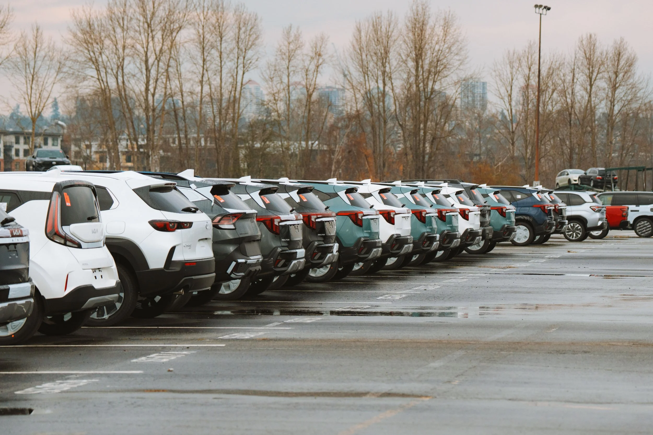A parking lot filled with various white and dark-colored SUVs and vehicles, with bare trees and city buildings in the background on a cloudy day.