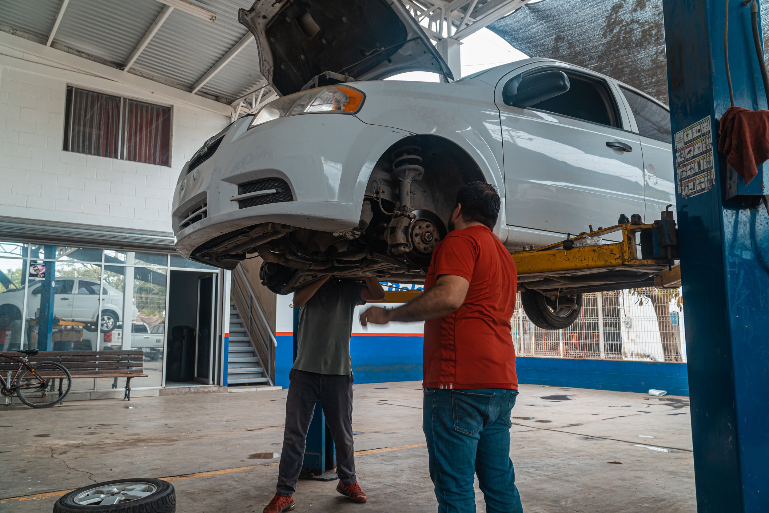 Two mechanics are working on a white car that is lifted on a hydraulic lift inside an auto repair shop. One mechanic is looking underneath the car while the other is standing nearby, both focusing on the work.
