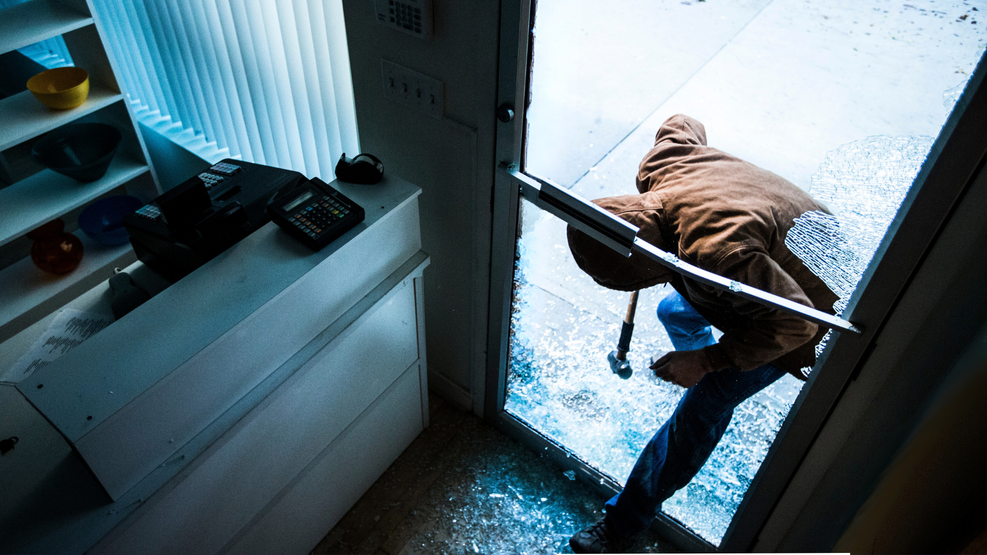 Person wearing a brown jacket and blue jeans breaking a glass door with a crowbar, glass shards on the ground, inside a building near a white shelf with bowls and a cash register.