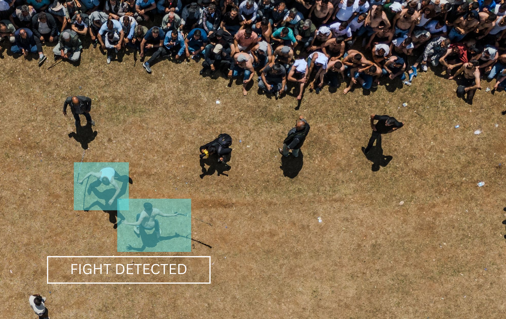 An aerial view of a large crowd of people gathered outdoors on a grassy area, with a few individuals standing apart from the crowd. A digital overlay shows a detected fight between two people, highlighted in blue boxes, with the label 'FIGHT DETECTED' in the lower left corner.
