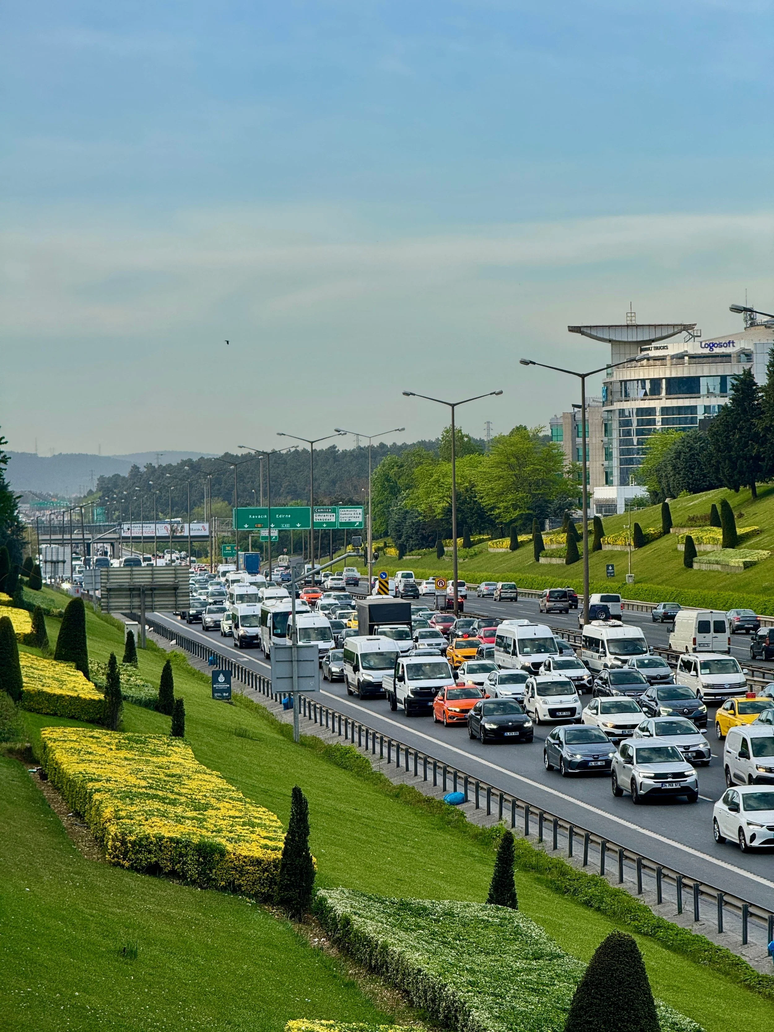 City highway with heavy traffic, surrounded by green landscaped areas and a modern building with signage, under a partly cloudy sky.