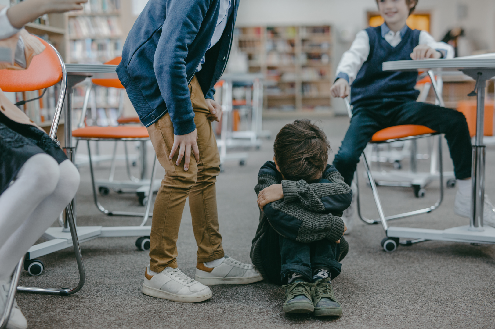 A boy sitting on the floor in a classroom, hugging his knees with his head down, while 3 other students taunt him, stand over him, and point at him.