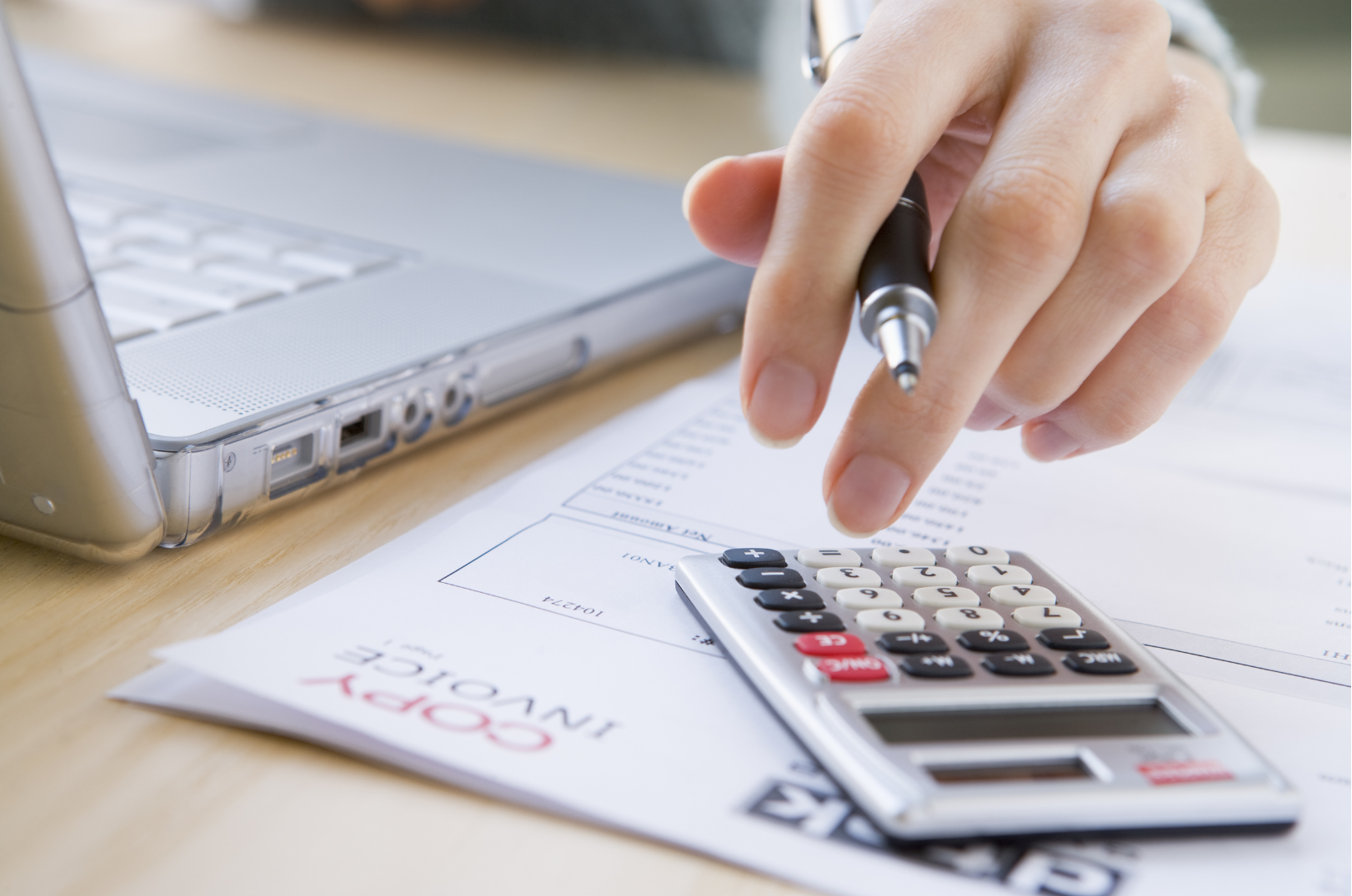 A person using a calculator, with a laptop computer and financial documents on a wooden desk.
