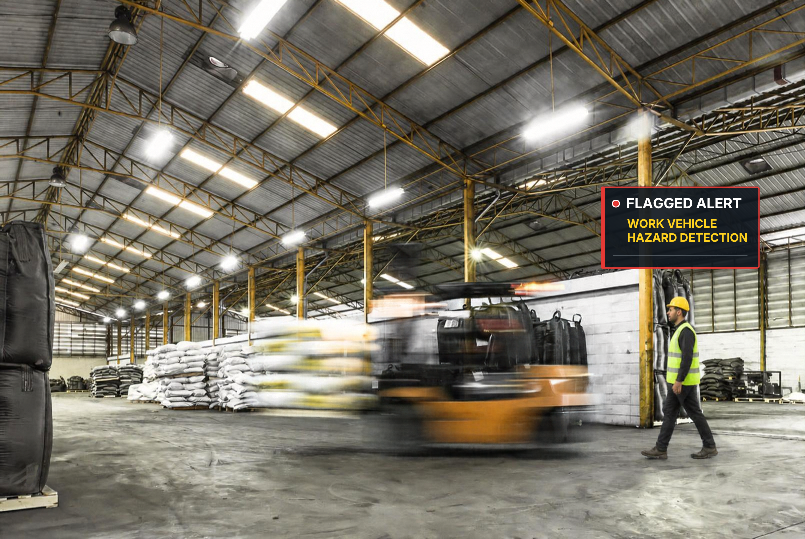 An industrial warehouse with metal roof and yellow support beams. A person wearing a yellow safety helmet and high visibility vest walks next to a wall with a digital alert sign reading 'Flagged Alert, Work Vehicle Hazard Detection' in the background. A blurred forklift moves across the floor, and there are stacks of materials including bags and padded items.