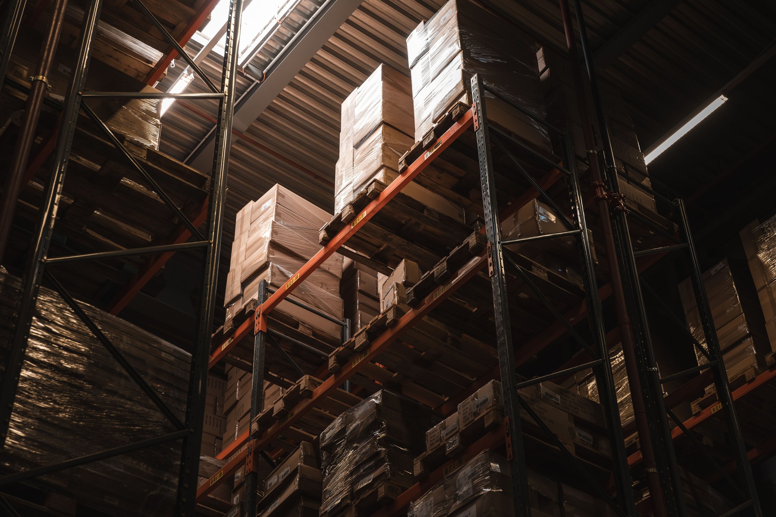 View of warehouse storage racks filled with stacked cardboard boxes wrapped in plastic, with high ceiling and metal support beams.