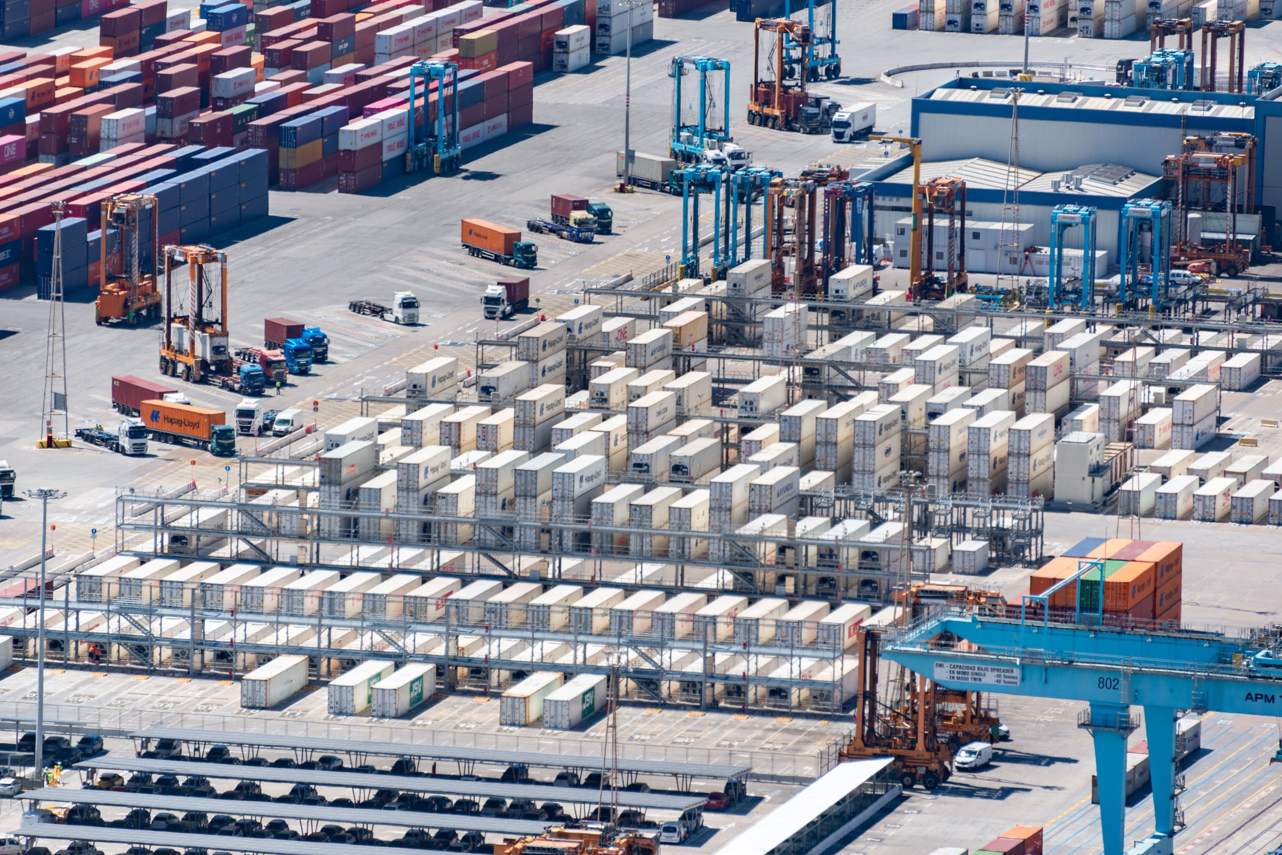 Aerial view of a large shipping port with numerous stacked containers, cranes, trucks, and heavy machinery.