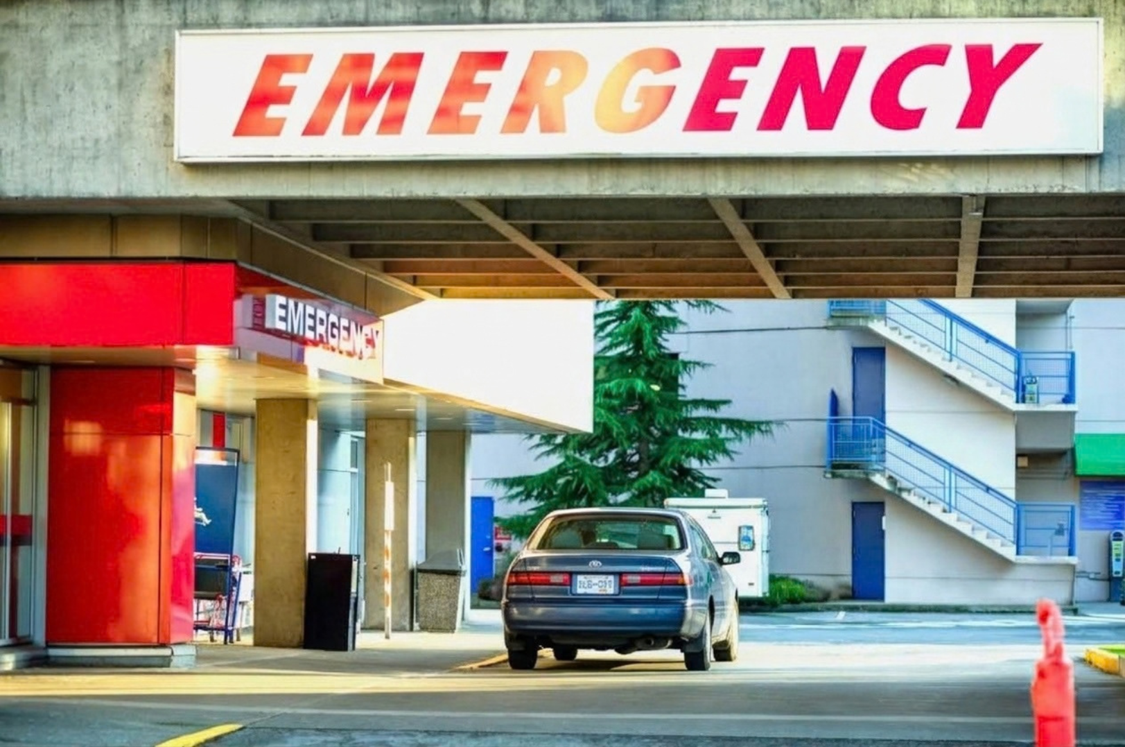 An emergency room entrance with a large sign that reads 'EMERGENCY.' A car is parked in front of the entrance that is reserved for emergency vehicles, and there is a blue staircase on the right side of the building.