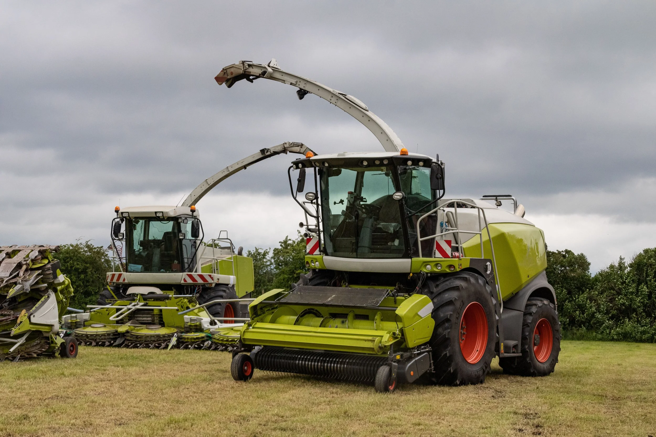 Two large green agricultural harvesters with black tires and orange accents, parked on grass under a cloudy sky.