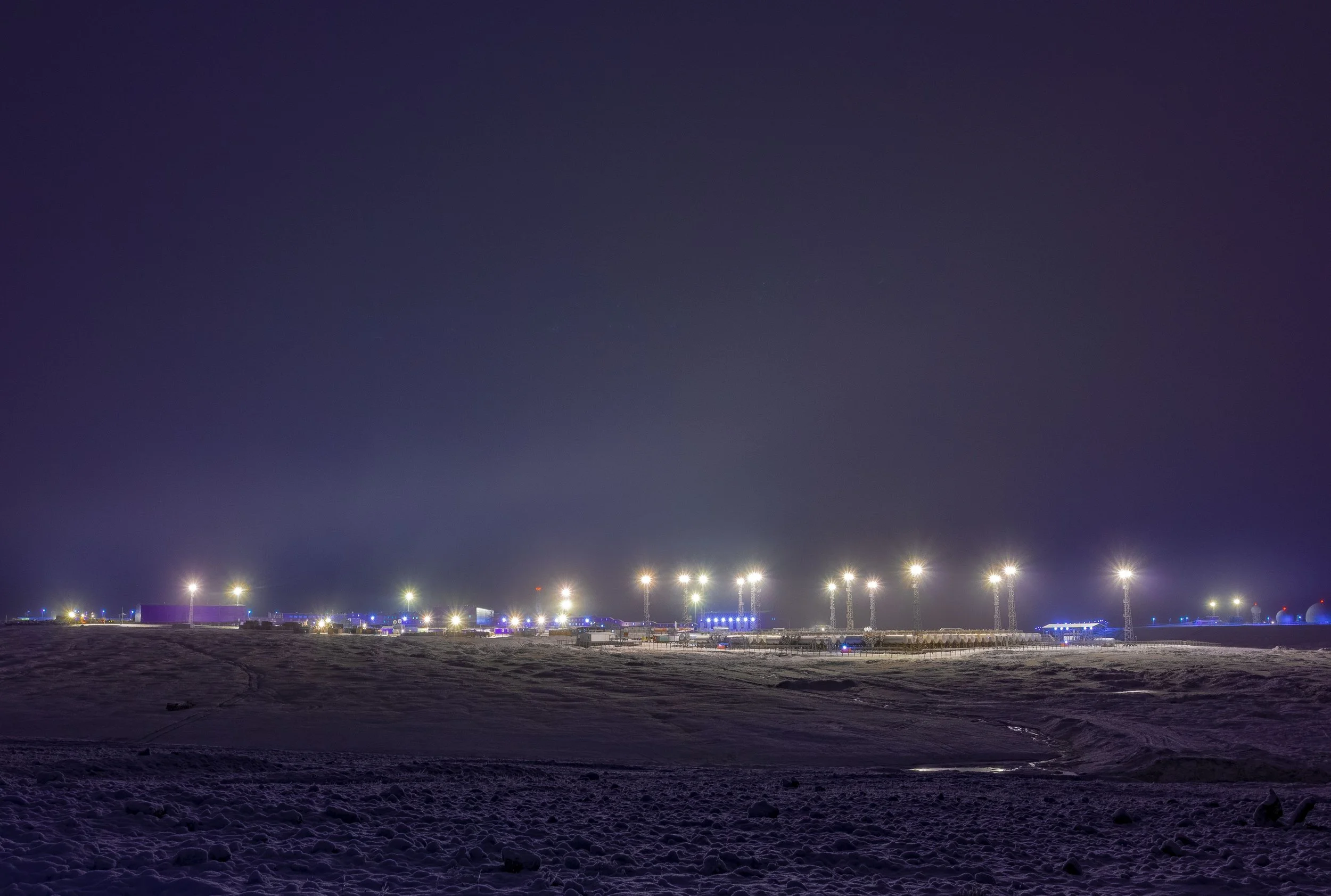 Nighttime view of an airport or industrial area with bright lights, several tall light poles, and a snow-covered ground in the foreground.