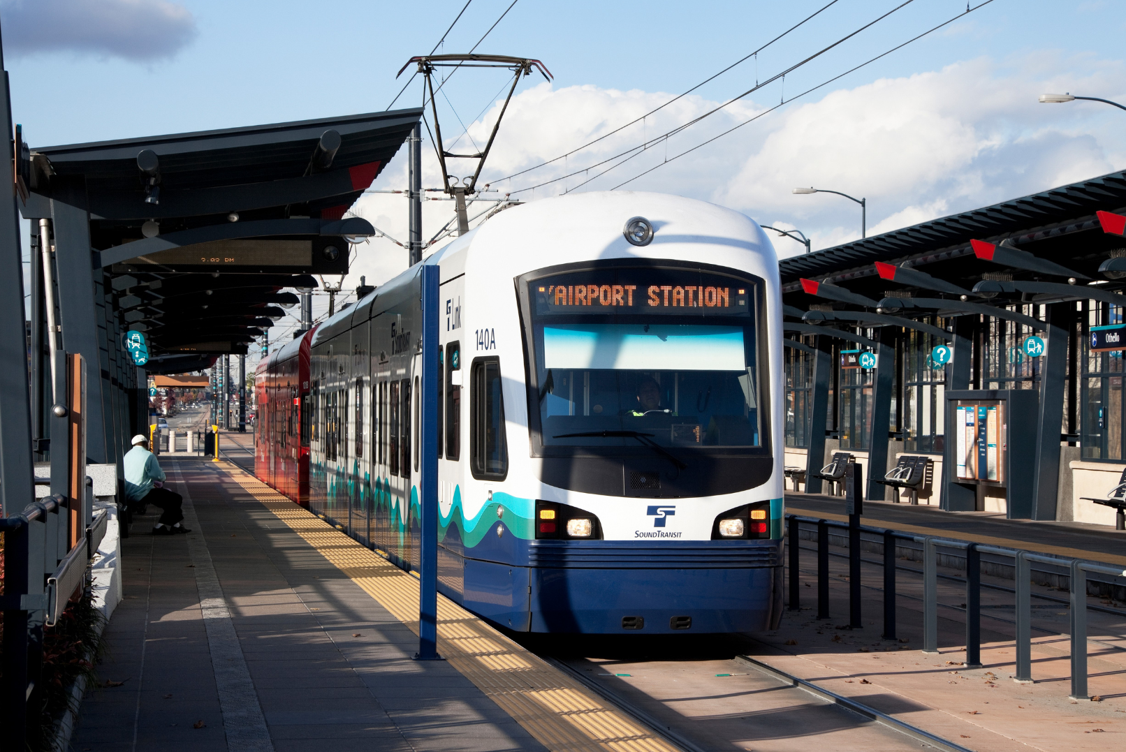 A modern light rail train at an outdoor station with blue and white colors, digital display indicating 'Airport Station,' and covered platform with benches and signs.