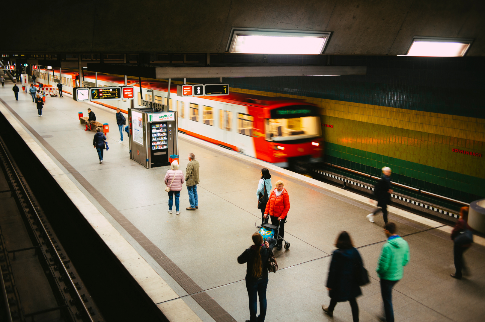 People waiting on a subway platform as a train arrives, with some sitting on benches and others walking or standing. The platform is inside a station with a colorful tiled wall in the background, and signs indicate the station name 'Rothenbach' and directions for train lines.