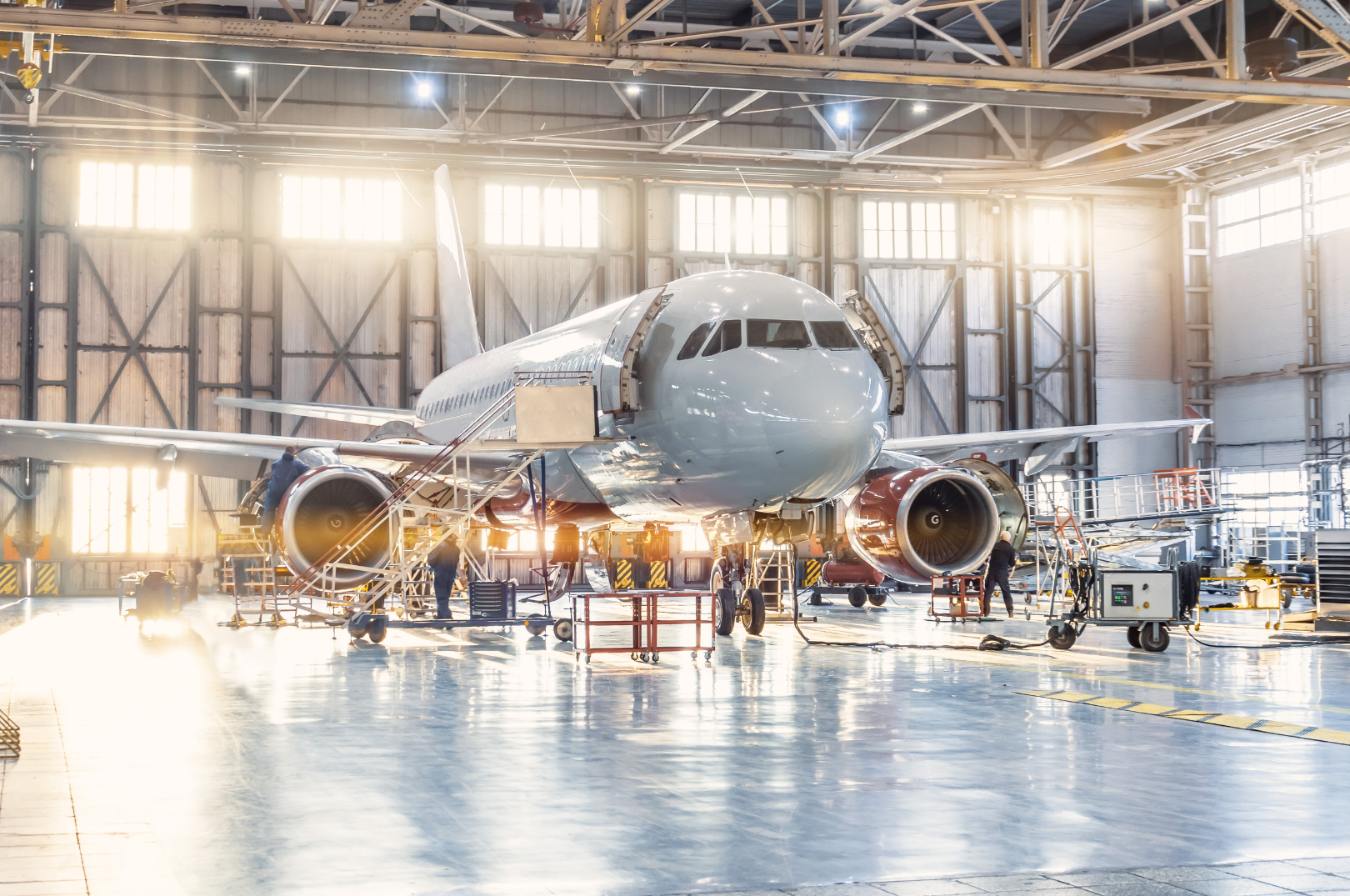 An airplane is inside an aircraft hangar with sunlight shining through the windows. The airplane is being serviced by workers, with equipment around it, and the hangar has a high ceiling with metal beams.