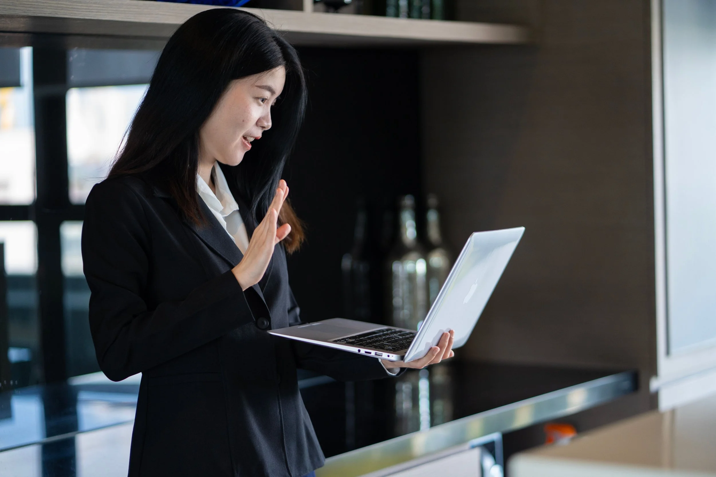 A woman in professional attire is holding a laptop and making a gesture with her hand, possibly a wave or stop signal, indoors near a shelf with books and bottles.