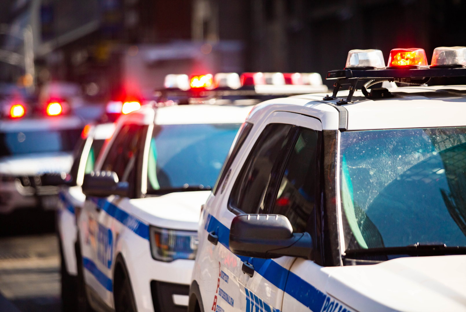 Multiple police cars with flashing lights parked on the street in daytime.