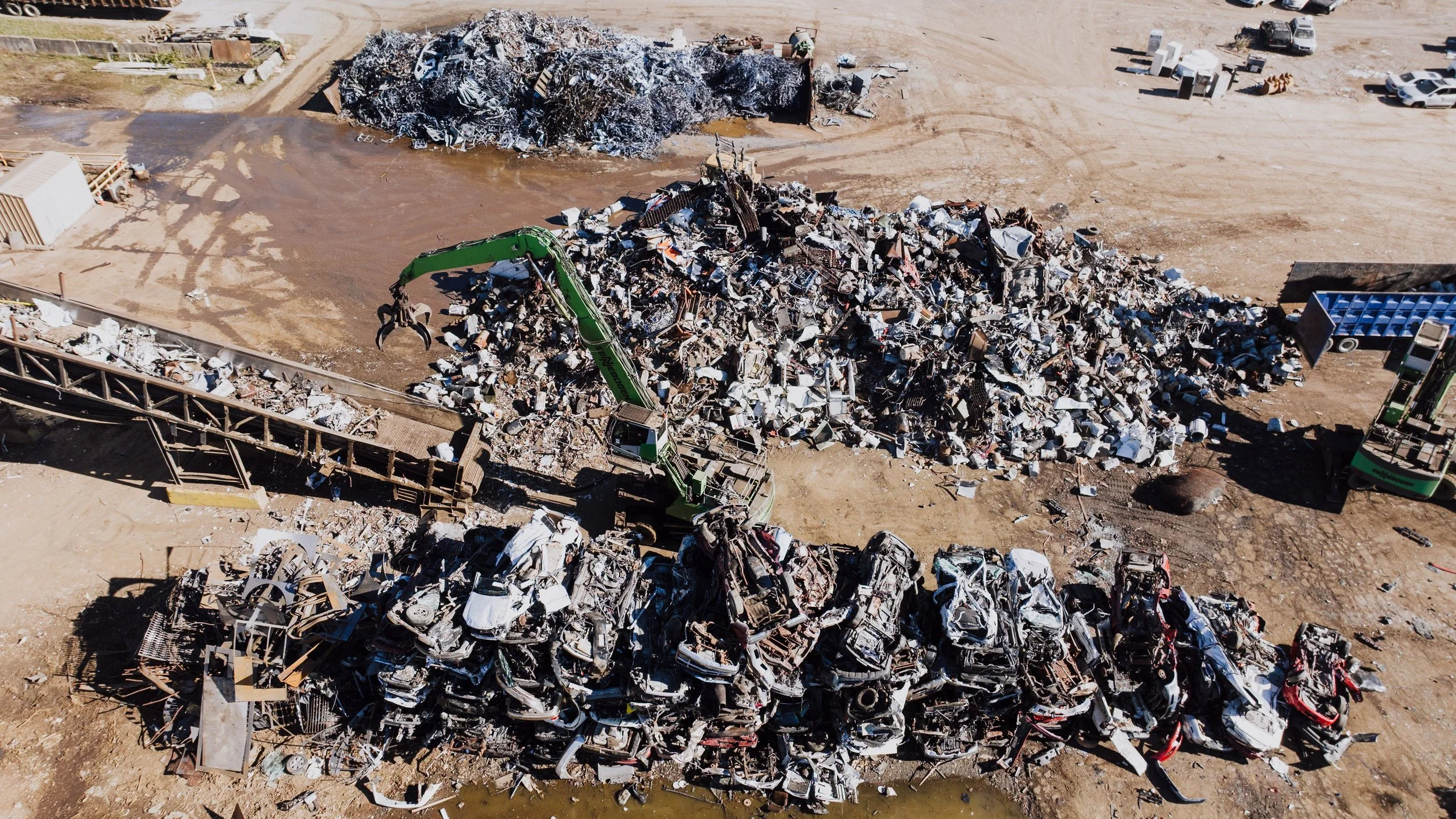 Aerial view of a metal yard with piles of crushed cars, machinery, and scattered debris on dirt ground.