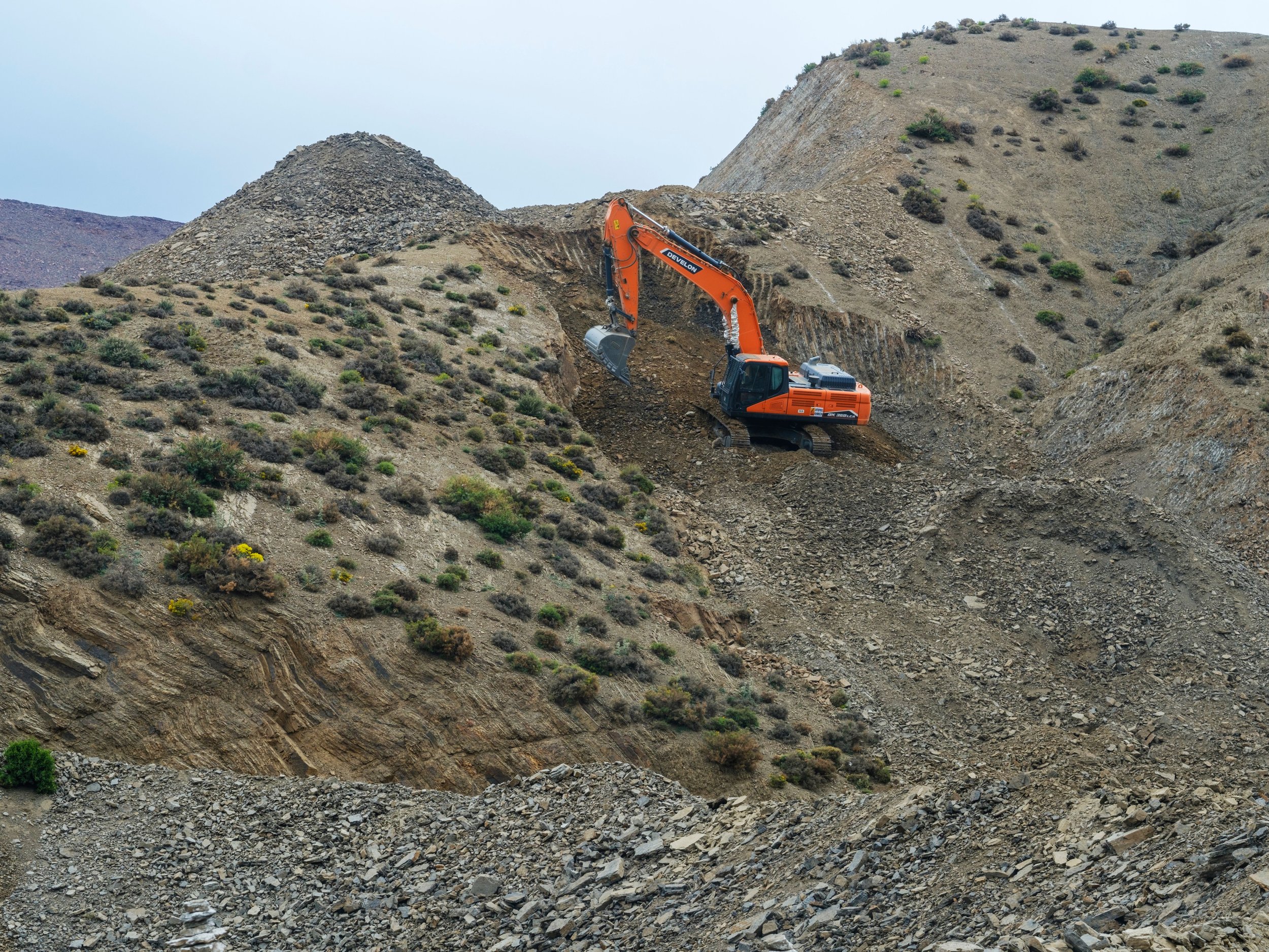 An orange excavator working on a mountain slope with sparse vegetation and rocky terrain.