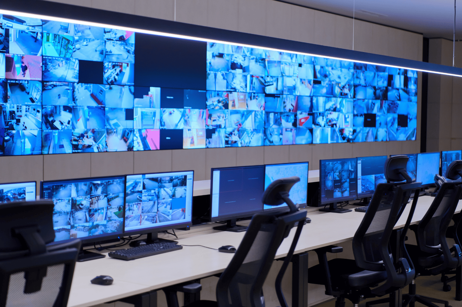 A control room with multiple computer monitors and a large video wall displaying surveillance camera feeds of a building.