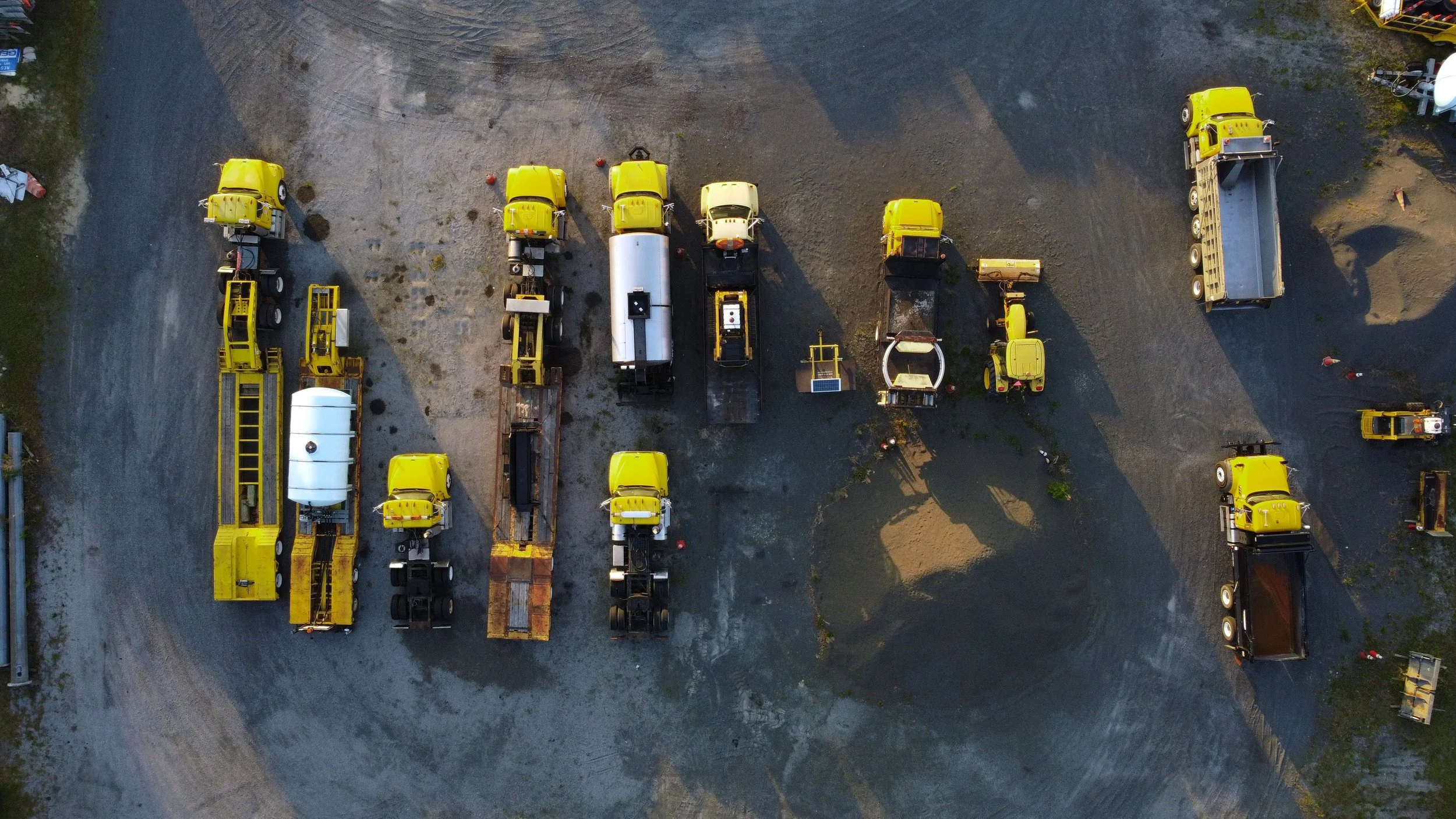 An aerial view of a gravel lot with several yellow construction vehicles and equipment parked in neat rows.
