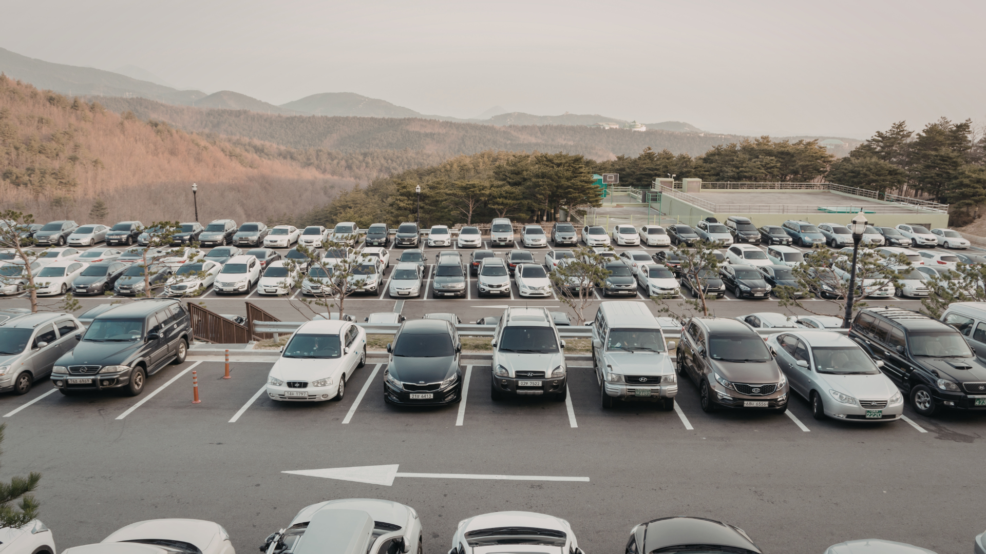 A parking lot with numerous parked cars in front of a scenic mountain landscape with trees and hills.