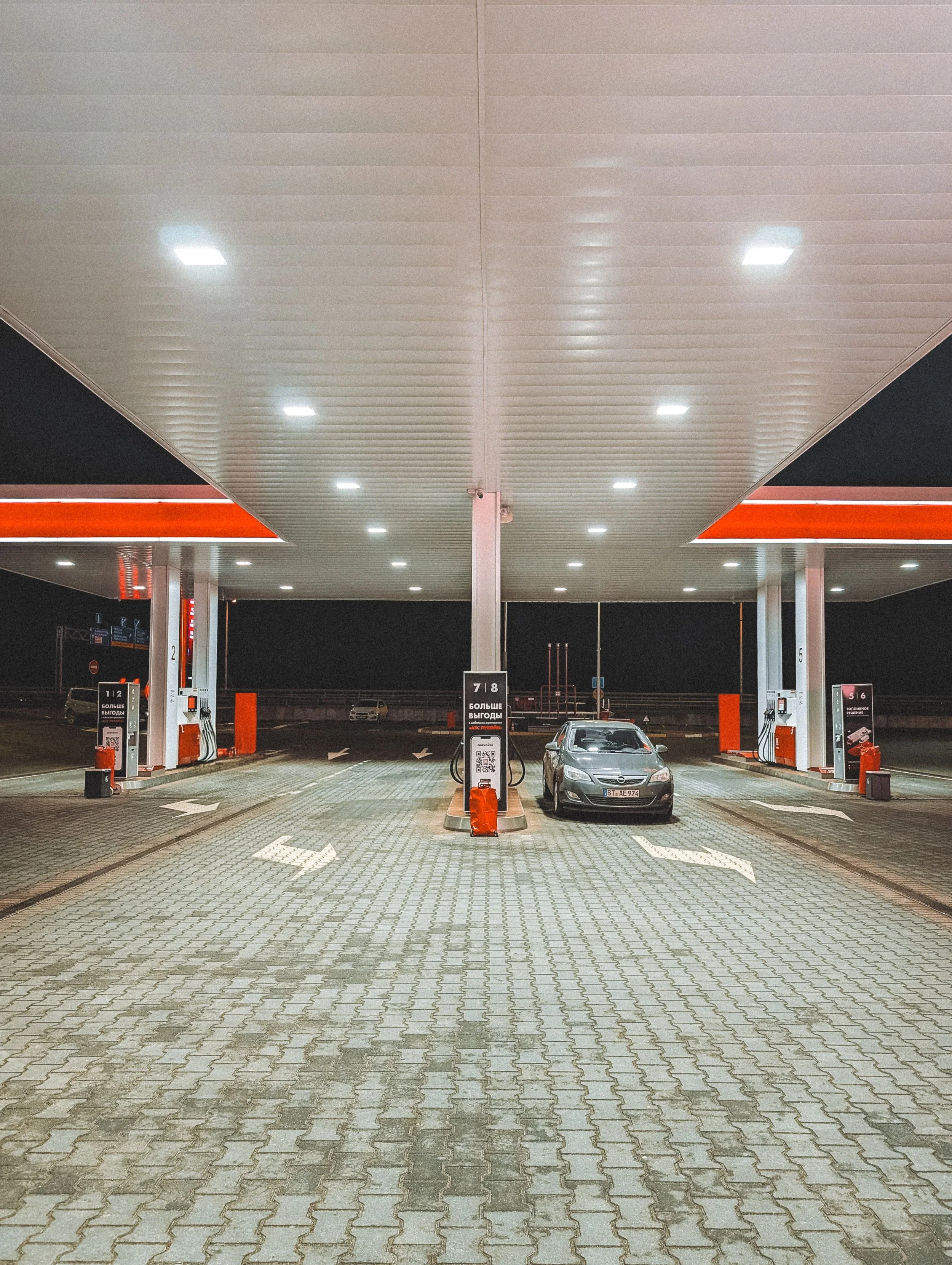 Empty gas station at night with two cars parked at pumps, illuminated canopy with lights, and paved ground with directional arrows.