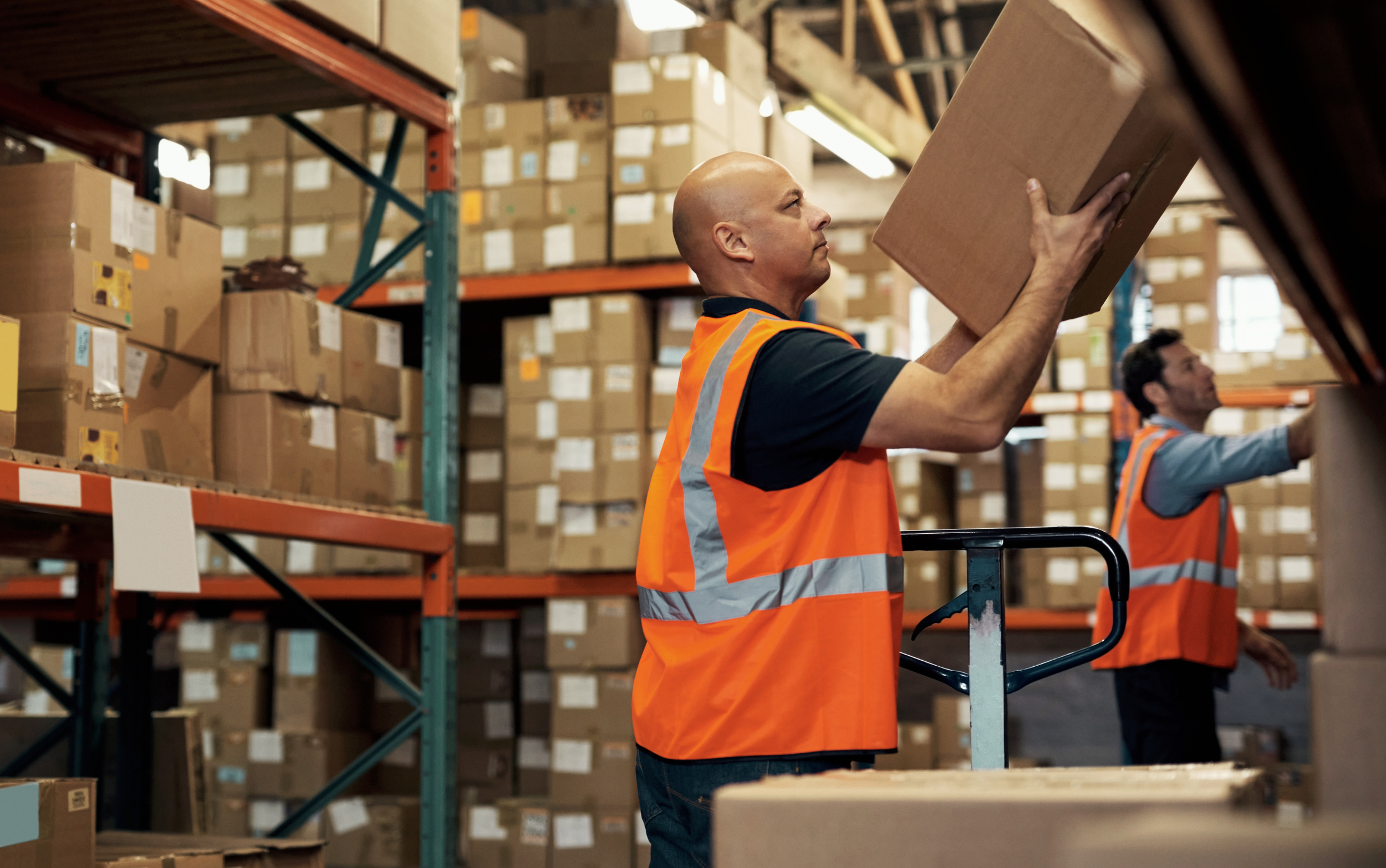 Man wearing an orange safety vest lifting a cardboard box in a warehouse filled with stacked boxes.