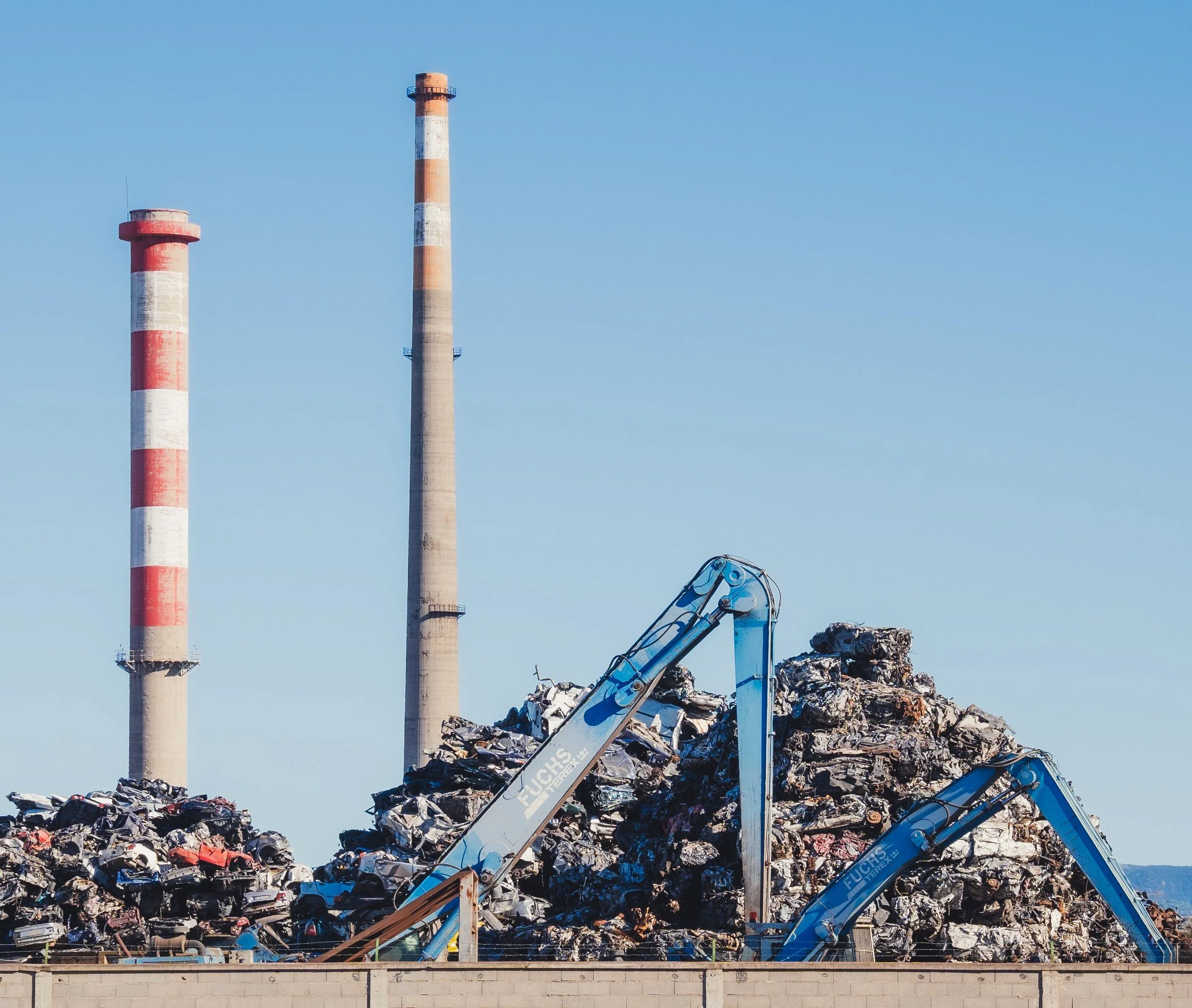 A demolition site with large piles of scrap metal and debris, a crane lifting metal, and three industrial smokestacks in the background against a clear blue sky.