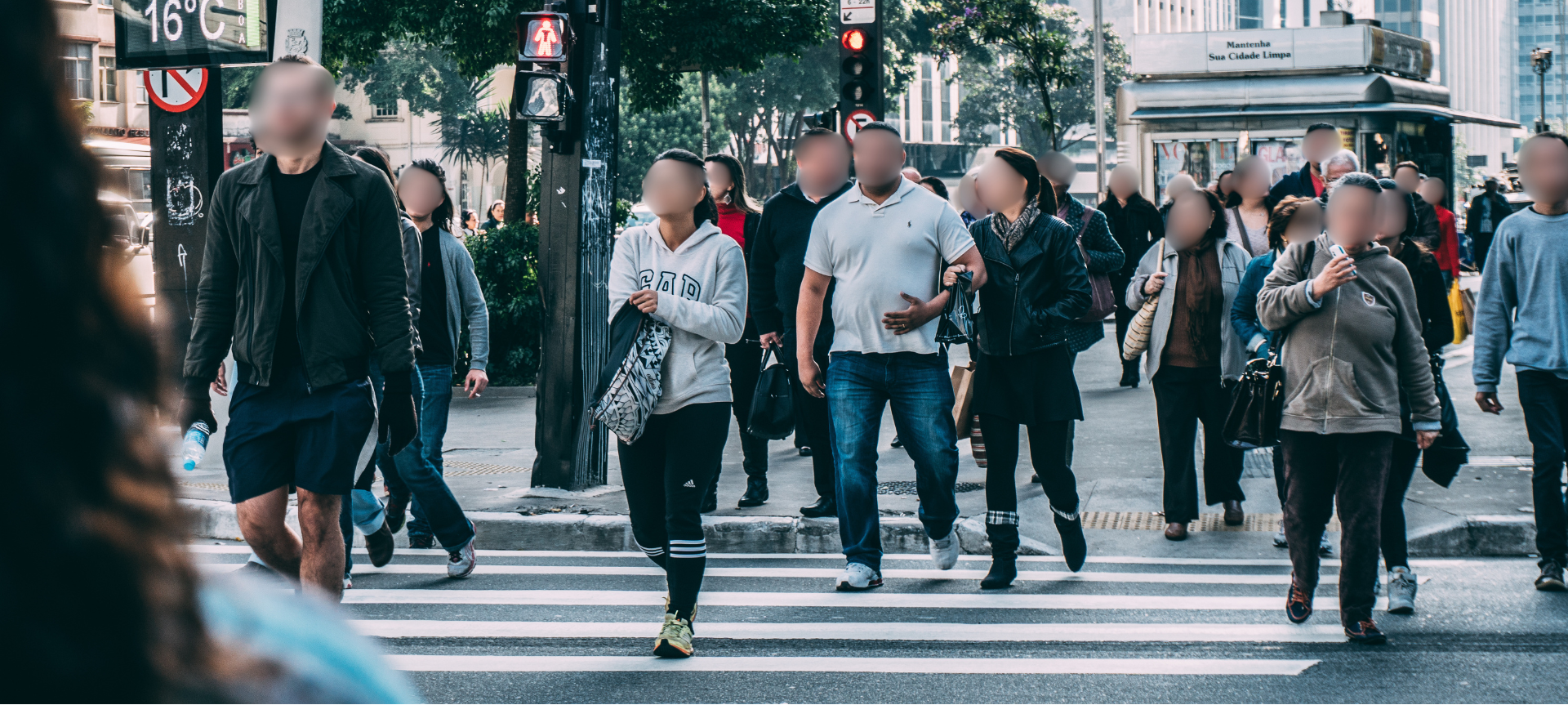 A busy city crosswalk with pedestrians crossing at a traffic light, some with shopping bags and backpacks, some using phones, with a city bus in the background.
