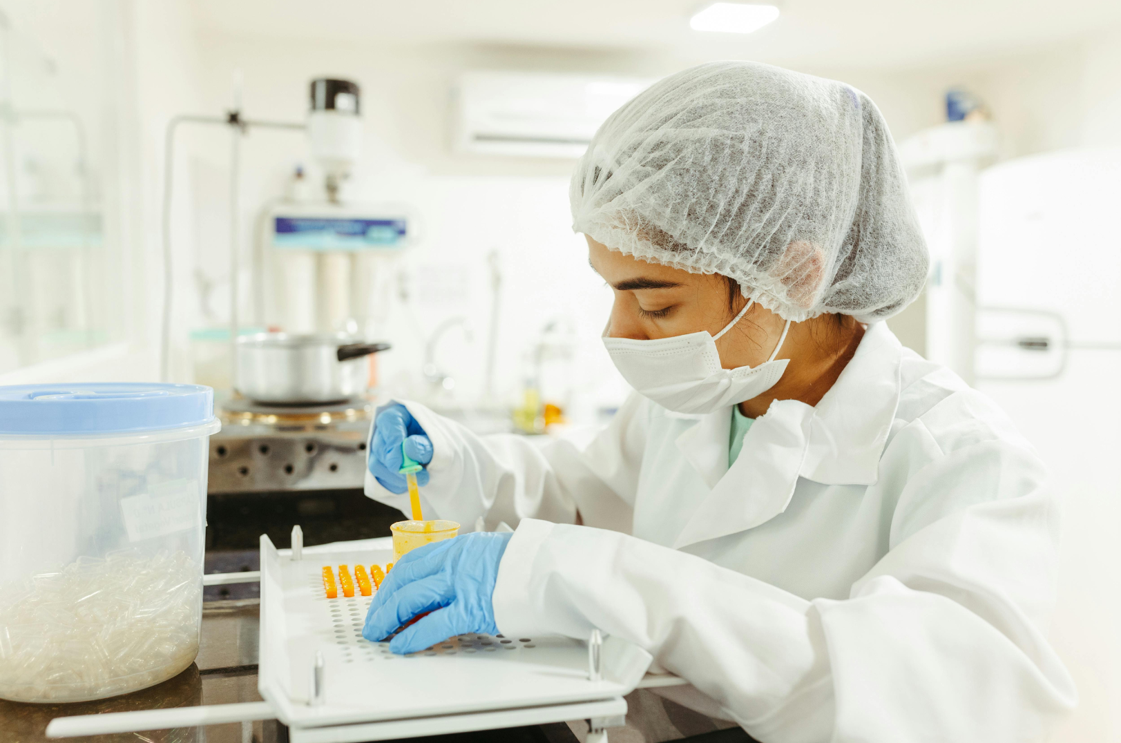 A scientist in a lab wearing a hairnet, face mask, gloves, and a white lab coat, working with test tubes and laboratory equipment.