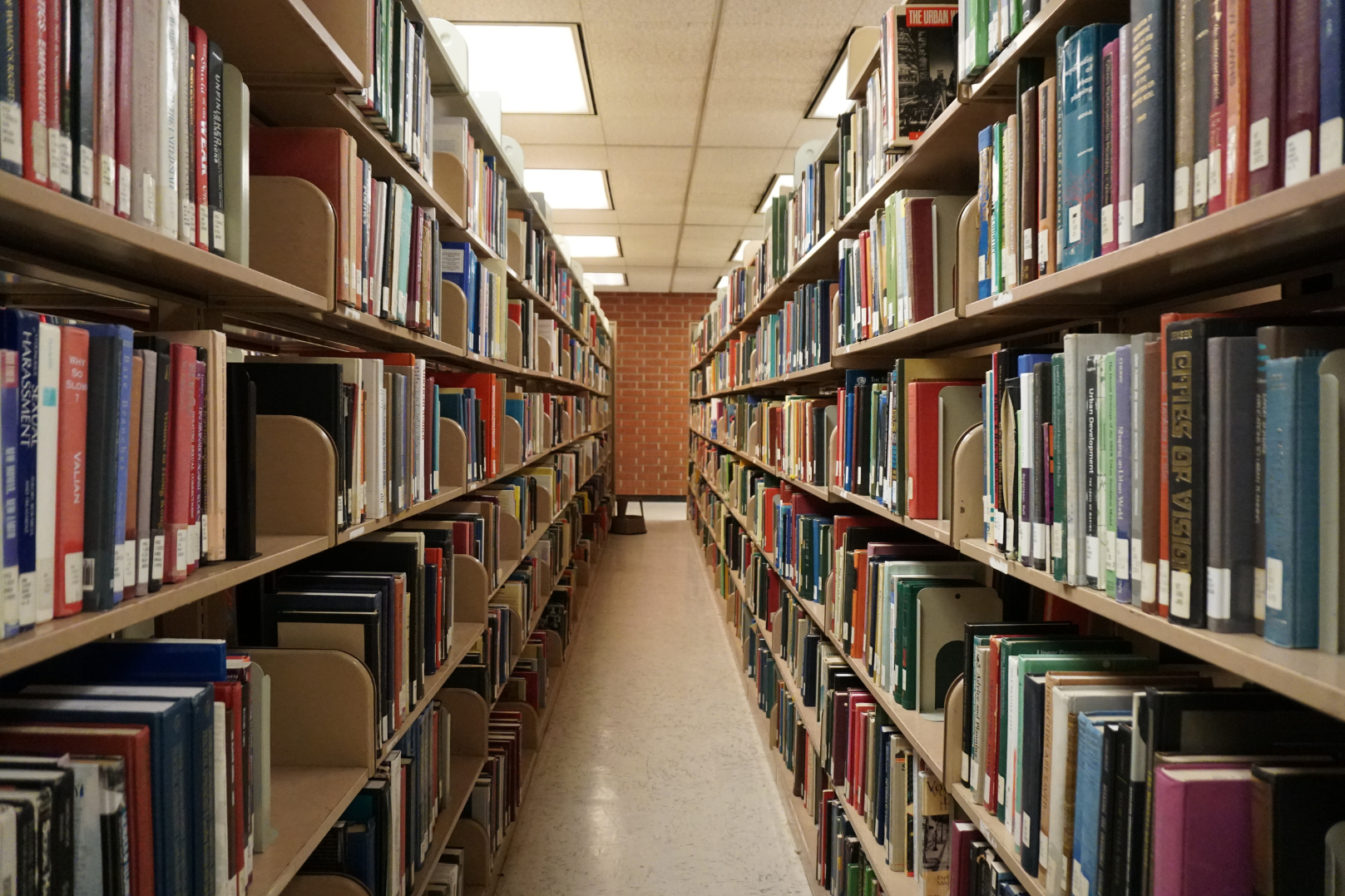 A narrow aisle in a library with brick wall at the end, bookshelves filled with books on both sides, and a small stool near the brick wall.