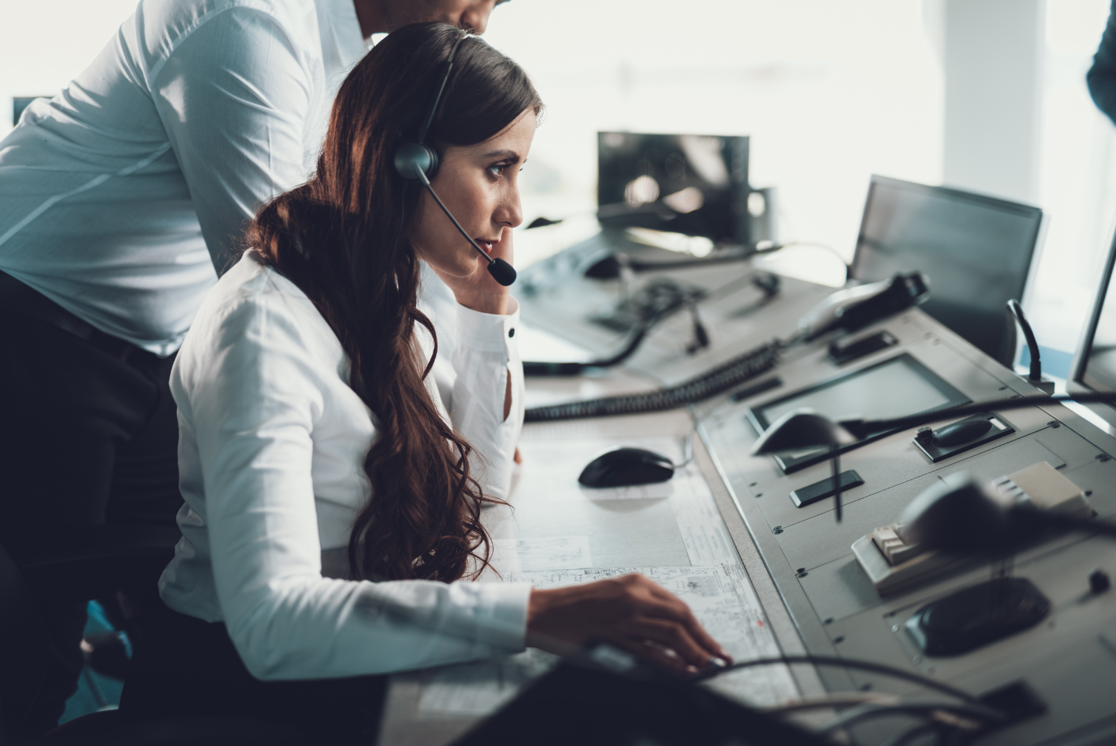 A woman wearing a headset sitting at a control panel, with a man leaning over her, in a professional office environment with multiple monitors and communication equipment.