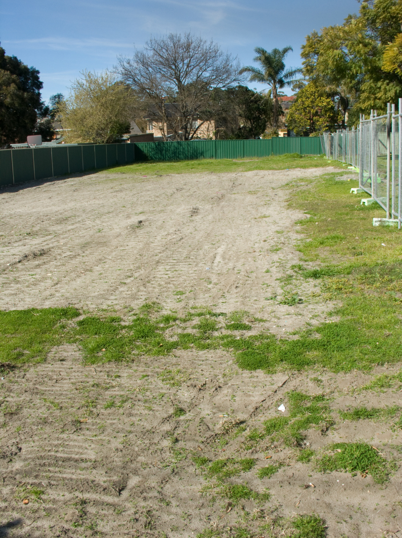 Empty fenced-in lot with patches of grass, tire tracks, and a row of trees and houses in the background under a clear blue sky.