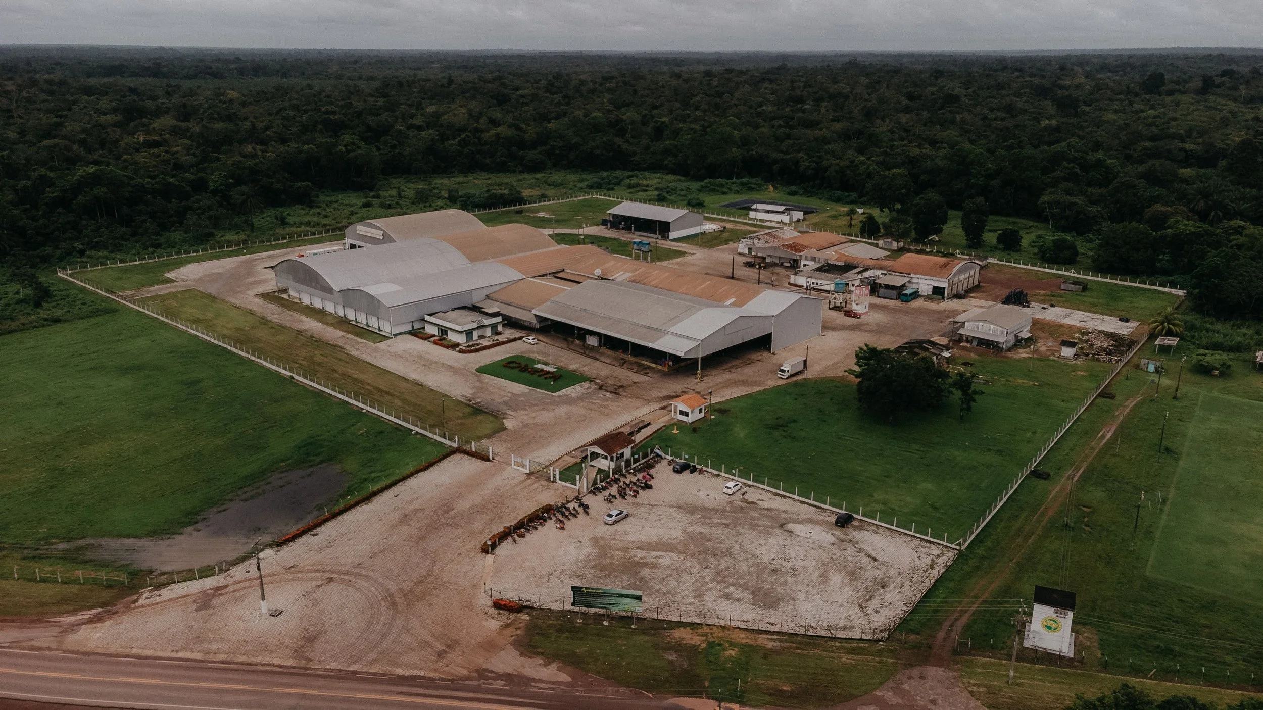 Aerial view of a rural industrial or commercial complex with multiple large buildings, surrounded by green fields and forested area, with a parking lot and a few vehicles nearby.