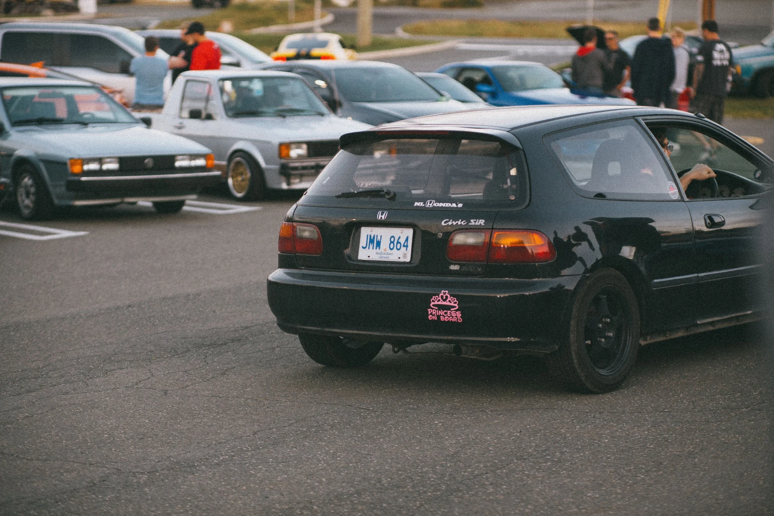 A parking lot with multiple cars and people standing around, with a focus on a black Honda Civic with a pink sticker that reads "Princess on Board".