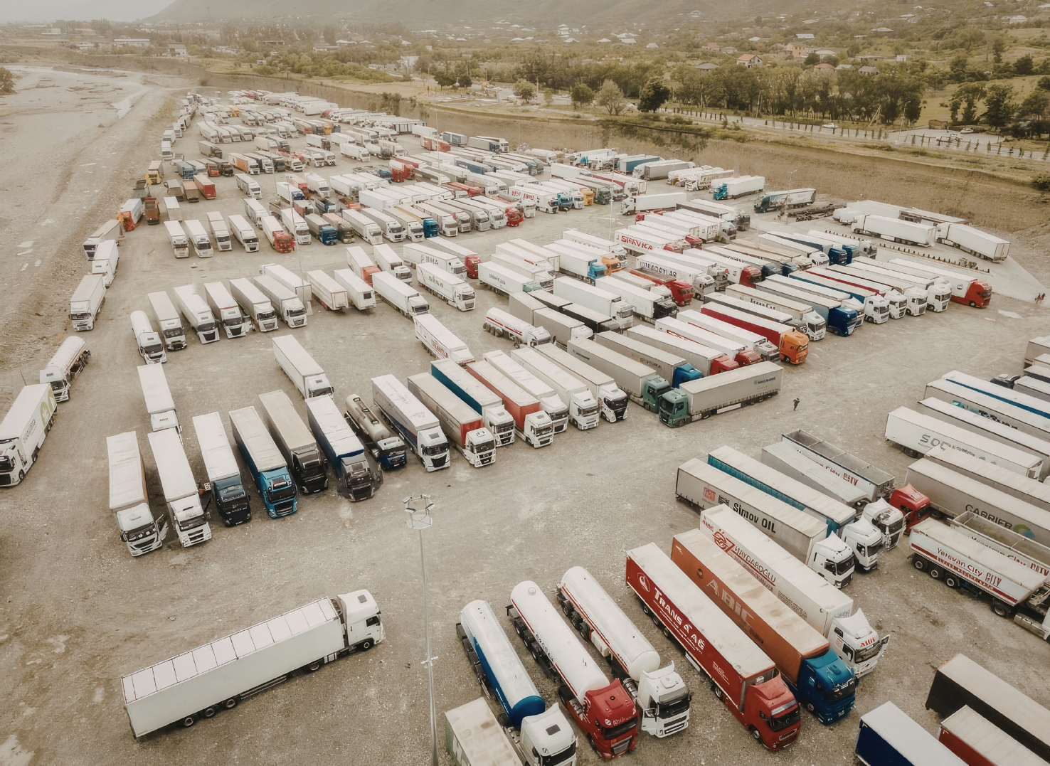 Aerial view of a large parking lot filled with numerous trucks and semi-trailers of various colors and brands, arranged in rows on a gravel surface.
