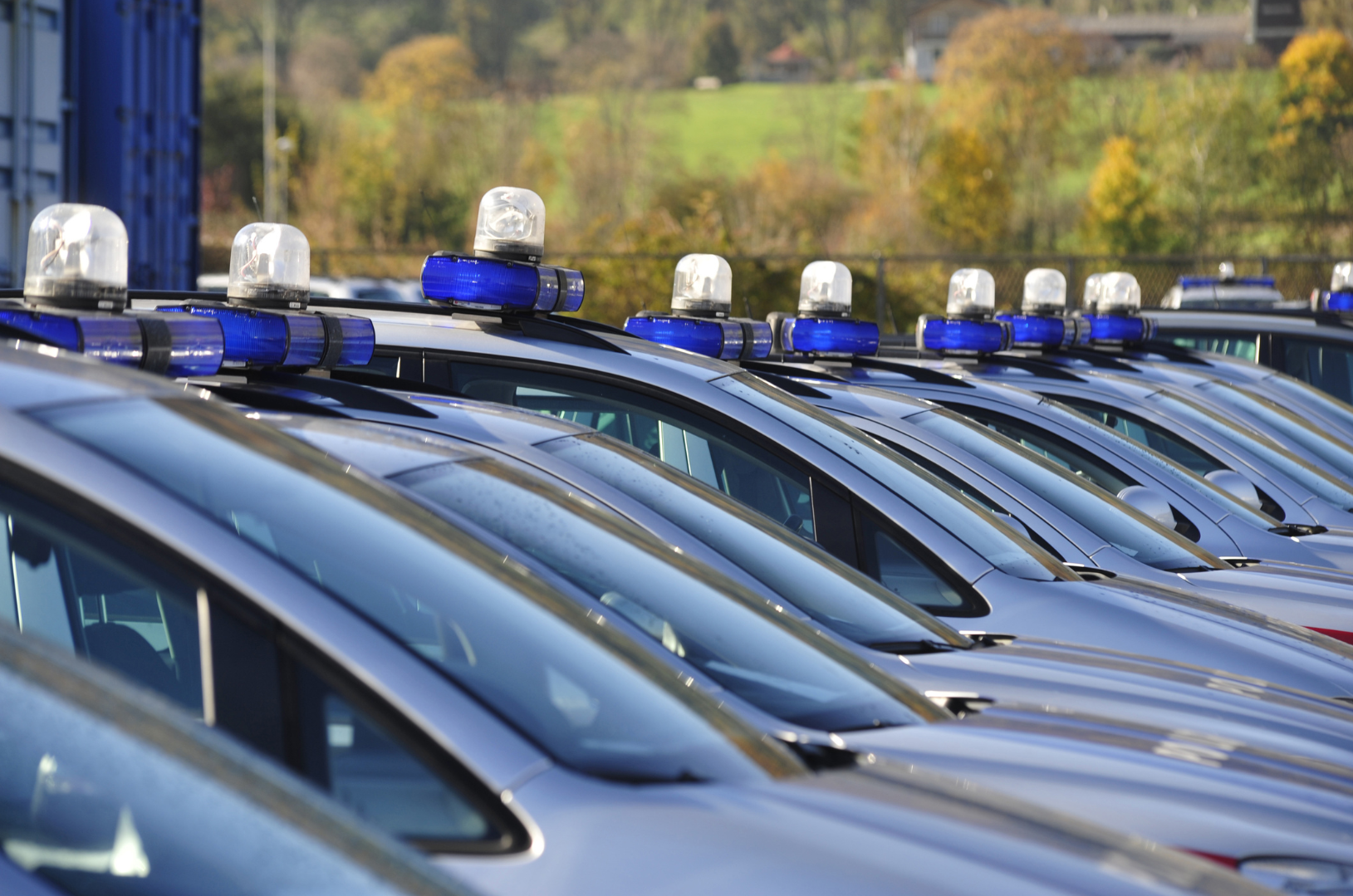 Multiple police cars lined up side by side with blue emergency lights on top, parked outdoors with trees and a hillside in the background.