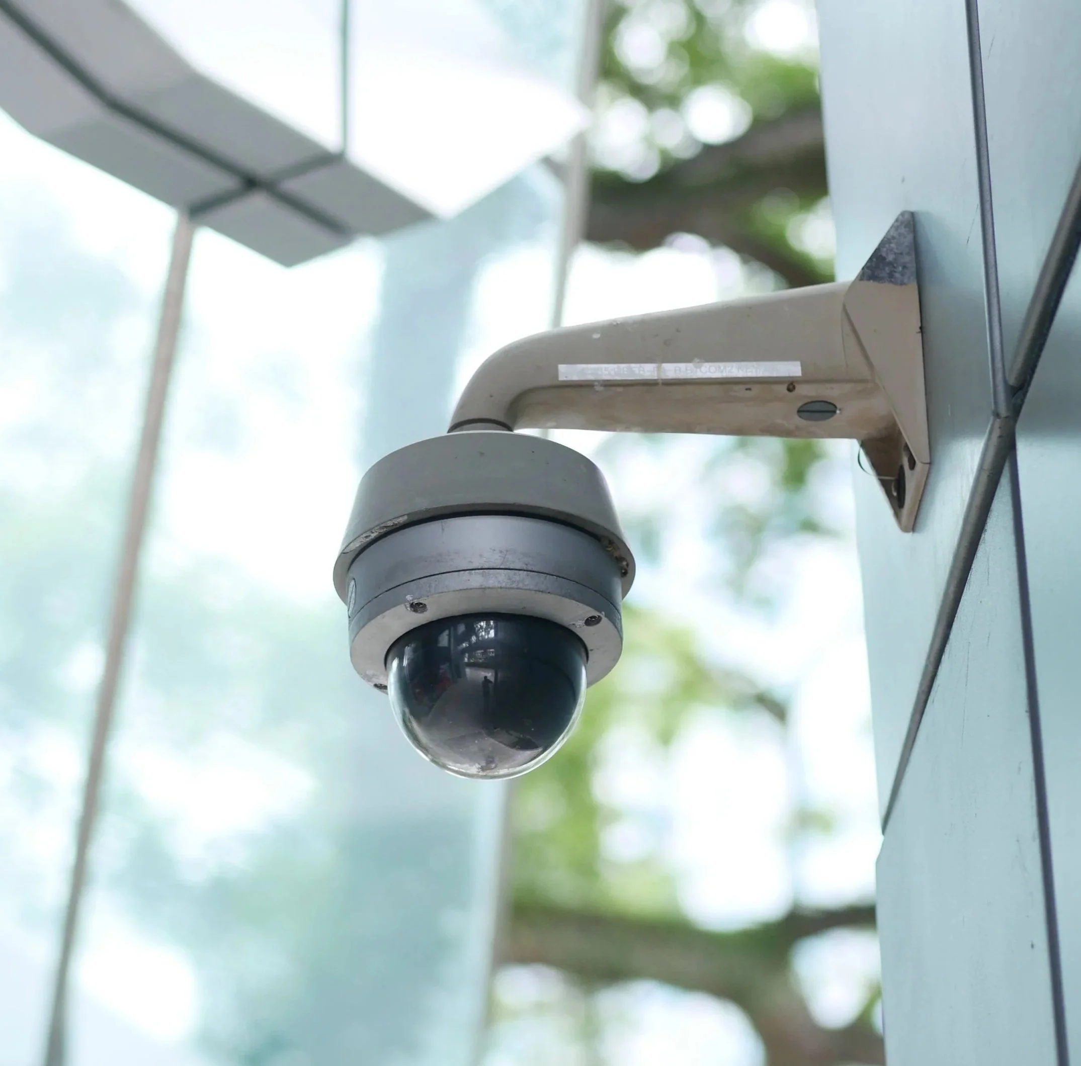 Close-up of a security camera mounted on a metal wall with blurred trees in the background.