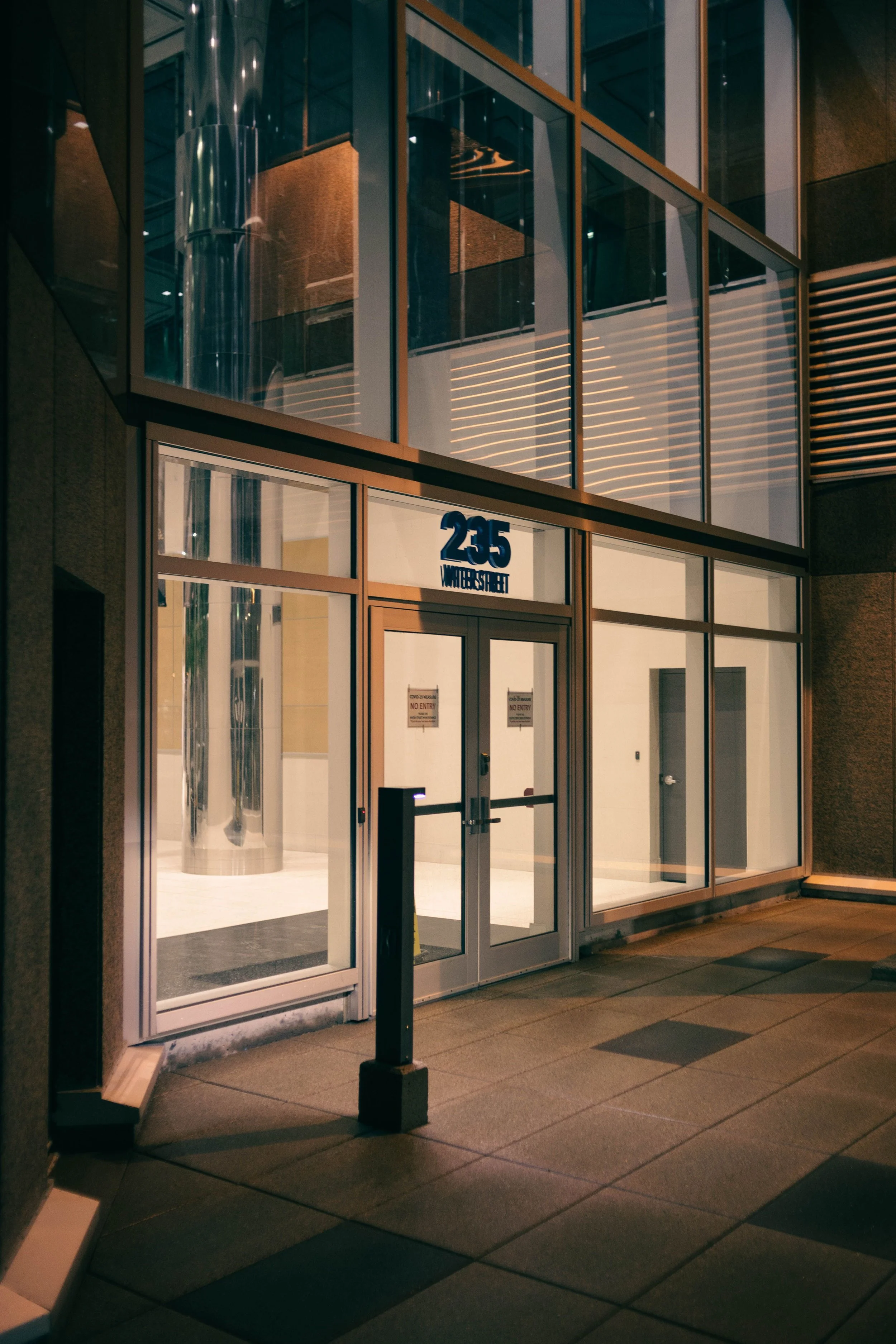 Nighttime view of the exterior entrance of a building with glass doors and large glass windows, displaying the street address '255 Warner Street.'