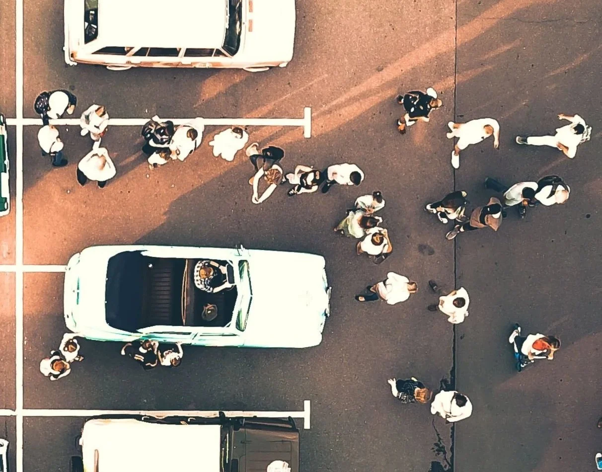 Top-down view of people walking in a parking lot with cars, some standing in line and others walking alone or in groups.