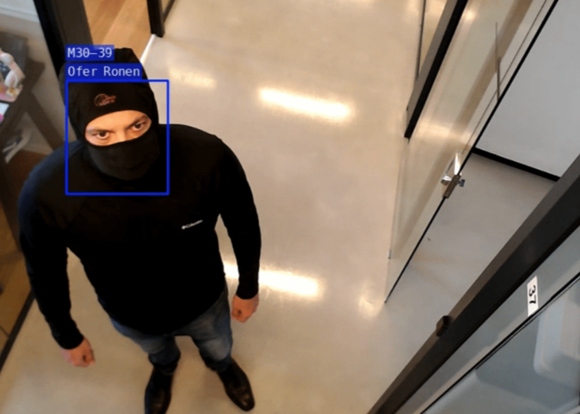 A man wearing a black mask, black beanie, and black clothing standing in an indoor setting, possibly a store or office, looking up at the camera.