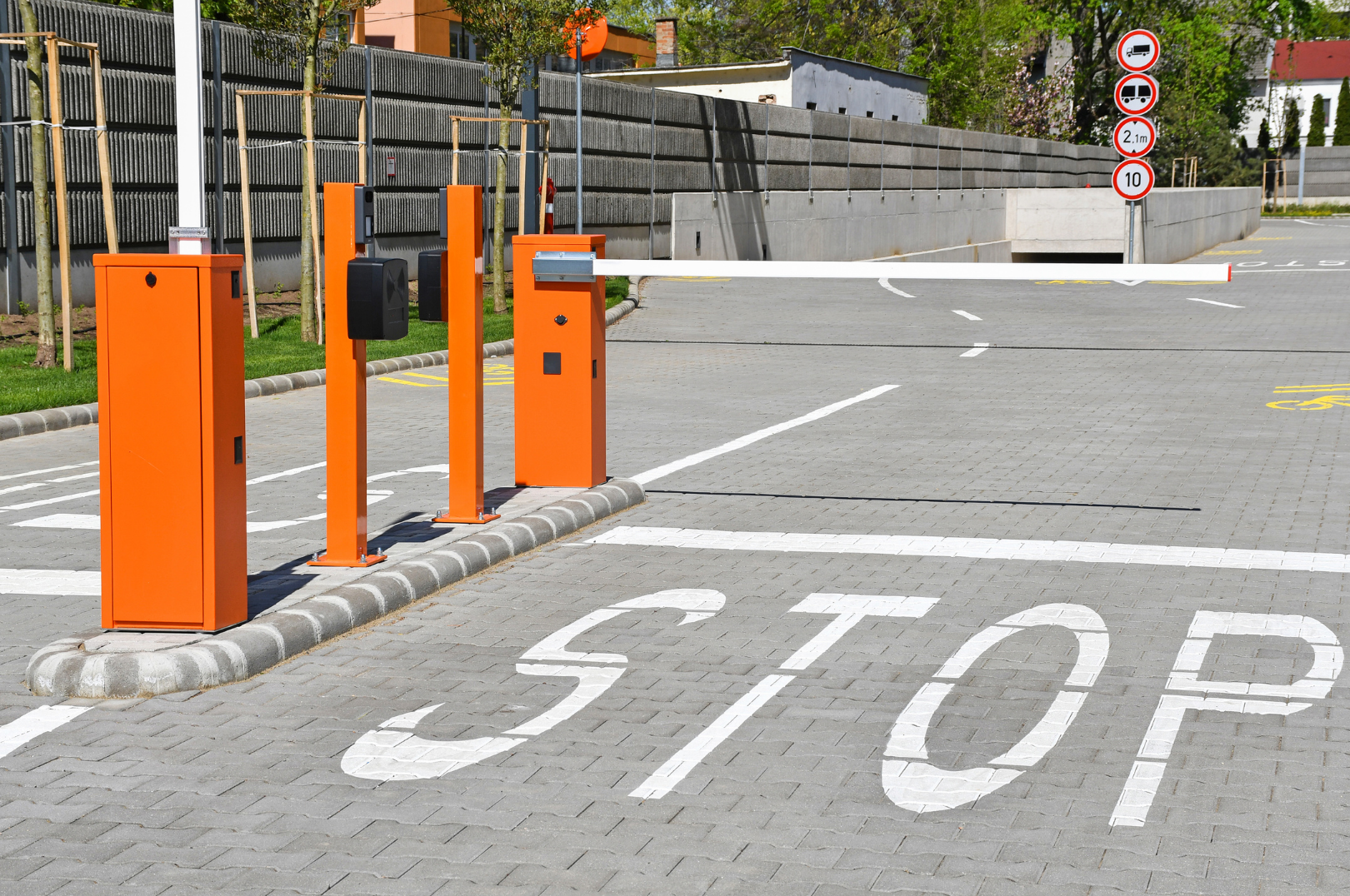 Parking lot entrance with bright orange automatic barrier gate and four nearby security boxes. There are multiple road signs on a post in the background. The ground is paved with grey bricks, and the word STOP is painted on the pavement in large white letters. Trees, buildings, and a grey fence are in the background.