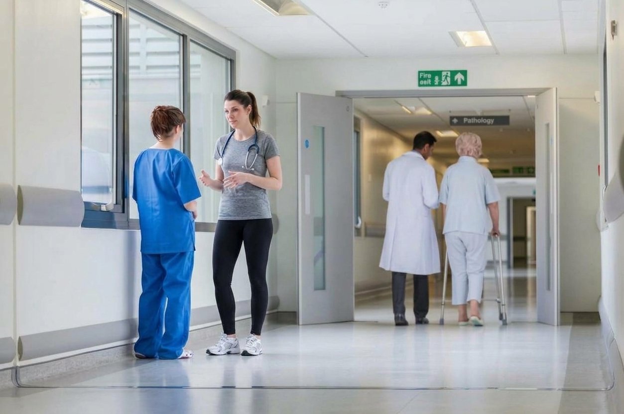 Hospital corridor with two healthcare workers talking near a window and two people with a walker and a medical white coat walking down the hall in the background.
