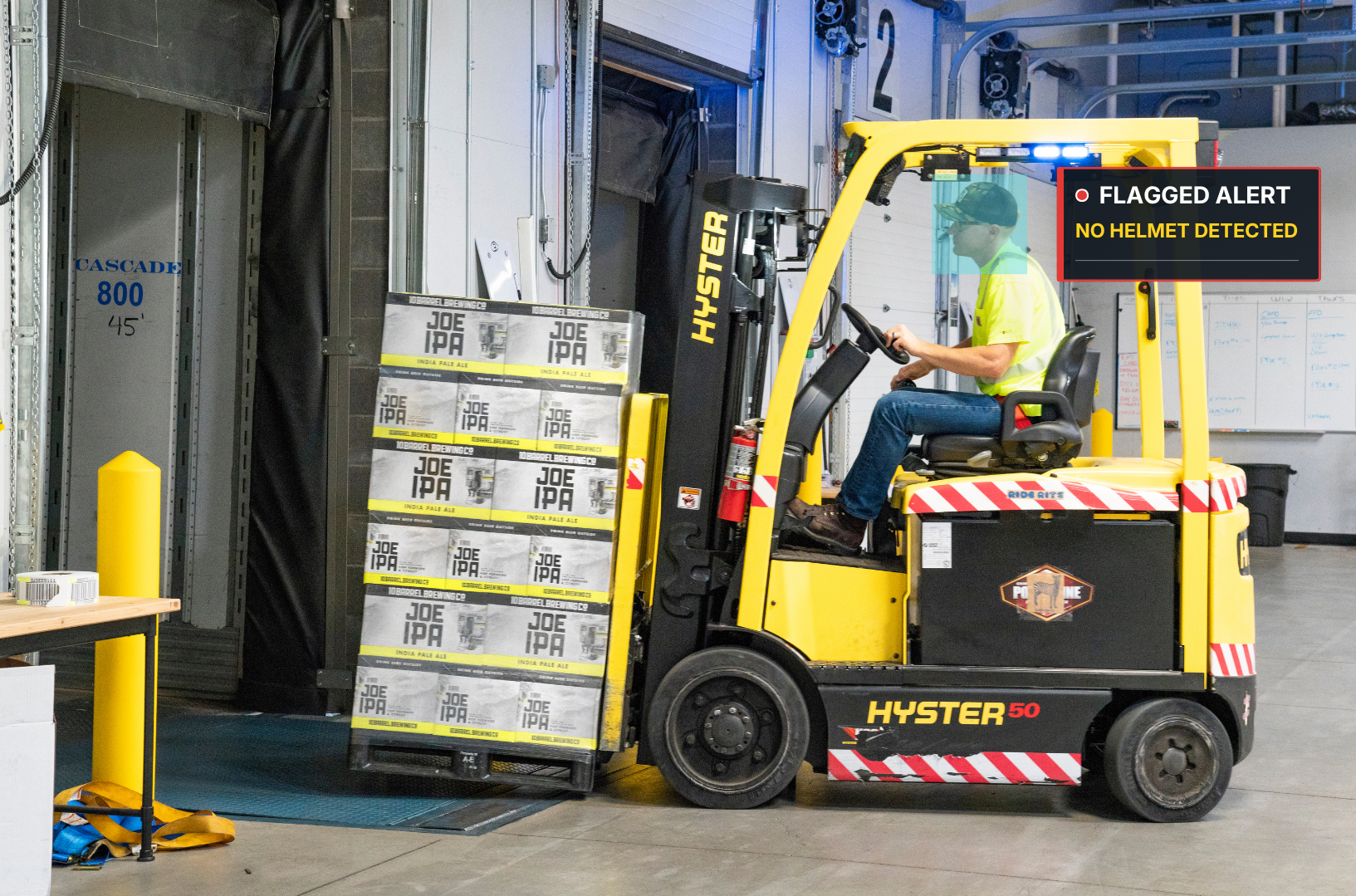 A forklift operator wearing a yellow safety shirt sitting on a yellow Hyster forklift in a warehouse, with a marked pallet of Joe IPA beer bottles in front. A digital alert reads 'Flagged Alert: No Helmet Detected' in the upper right.
